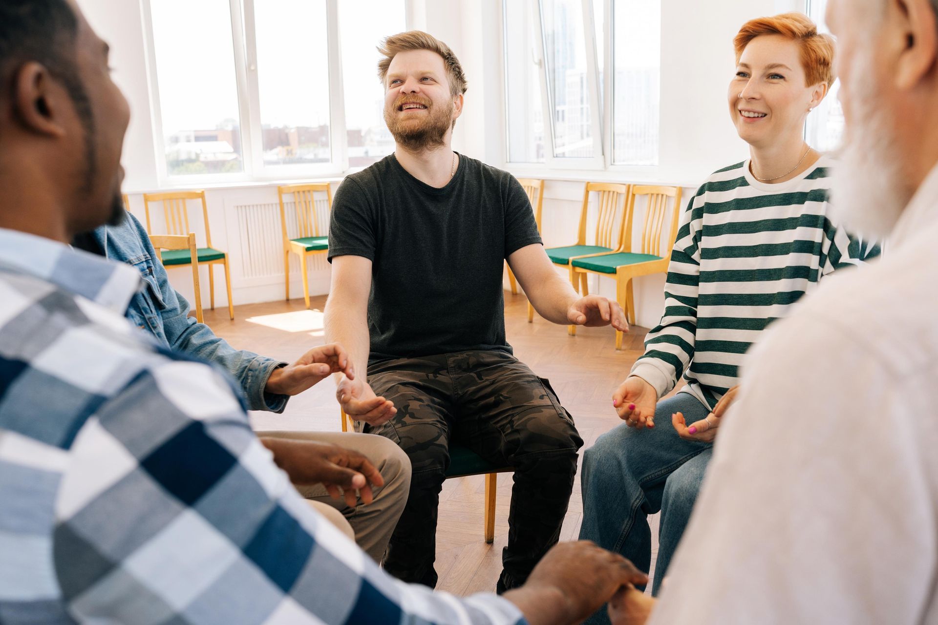 People in a circle, arms outstretched, engaged in a conversation in a room with windows and chairs.