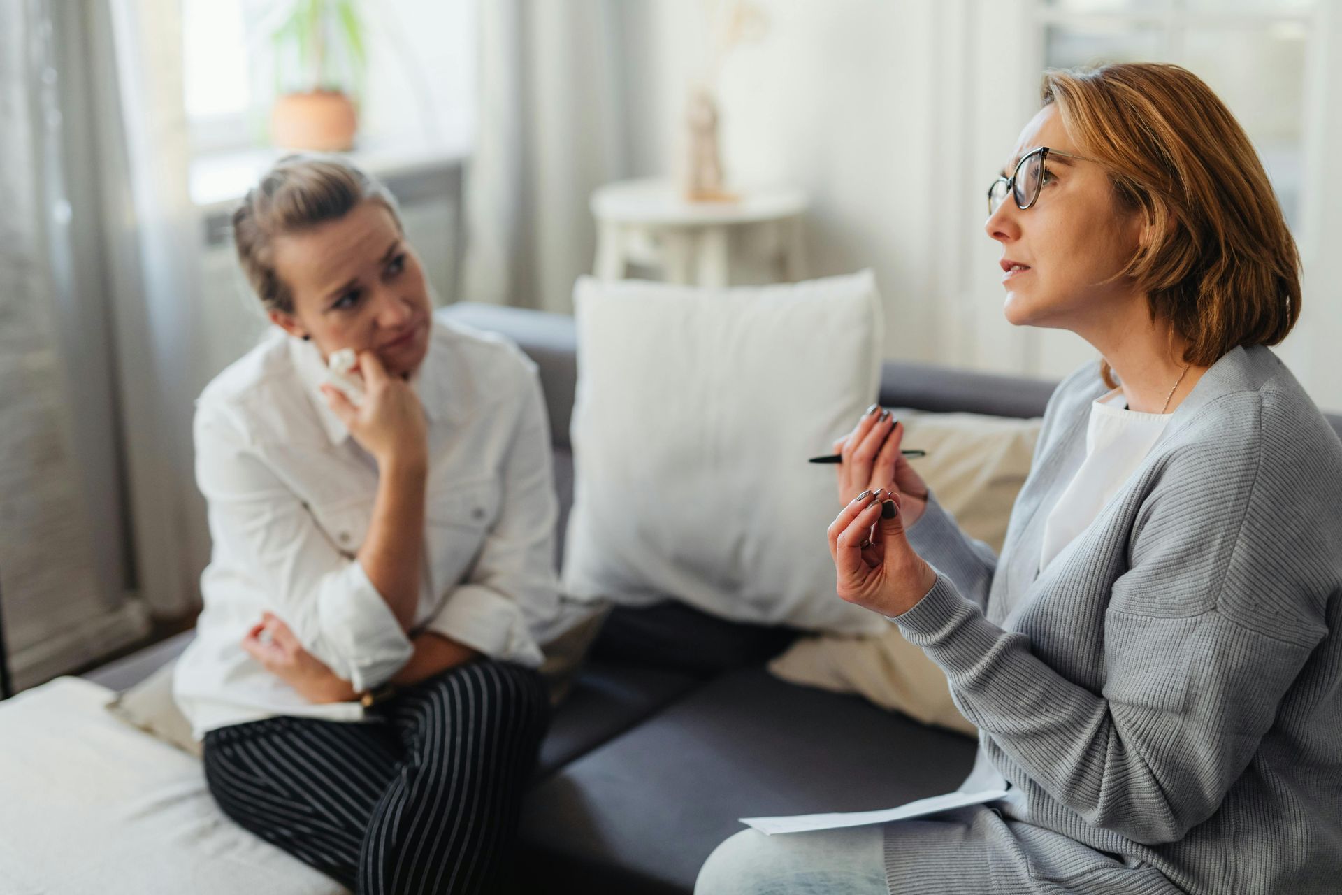 Two people talking on a couch in a bright living room, one holding a pen and the other listening attentively.