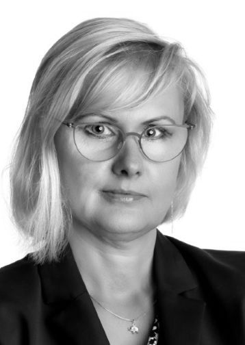 Žena s blond vlasy, brýlemi a sakem.