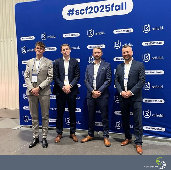 スーツを着た4人の男性が、イベントのブランドが入った青い背景の前でポーズをとっています。