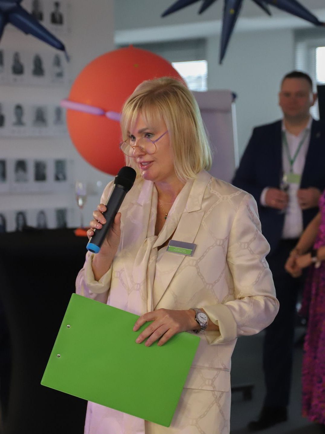 Woman speaking into a microphone, holding a green folder. She's in a light suit, in an event setting.