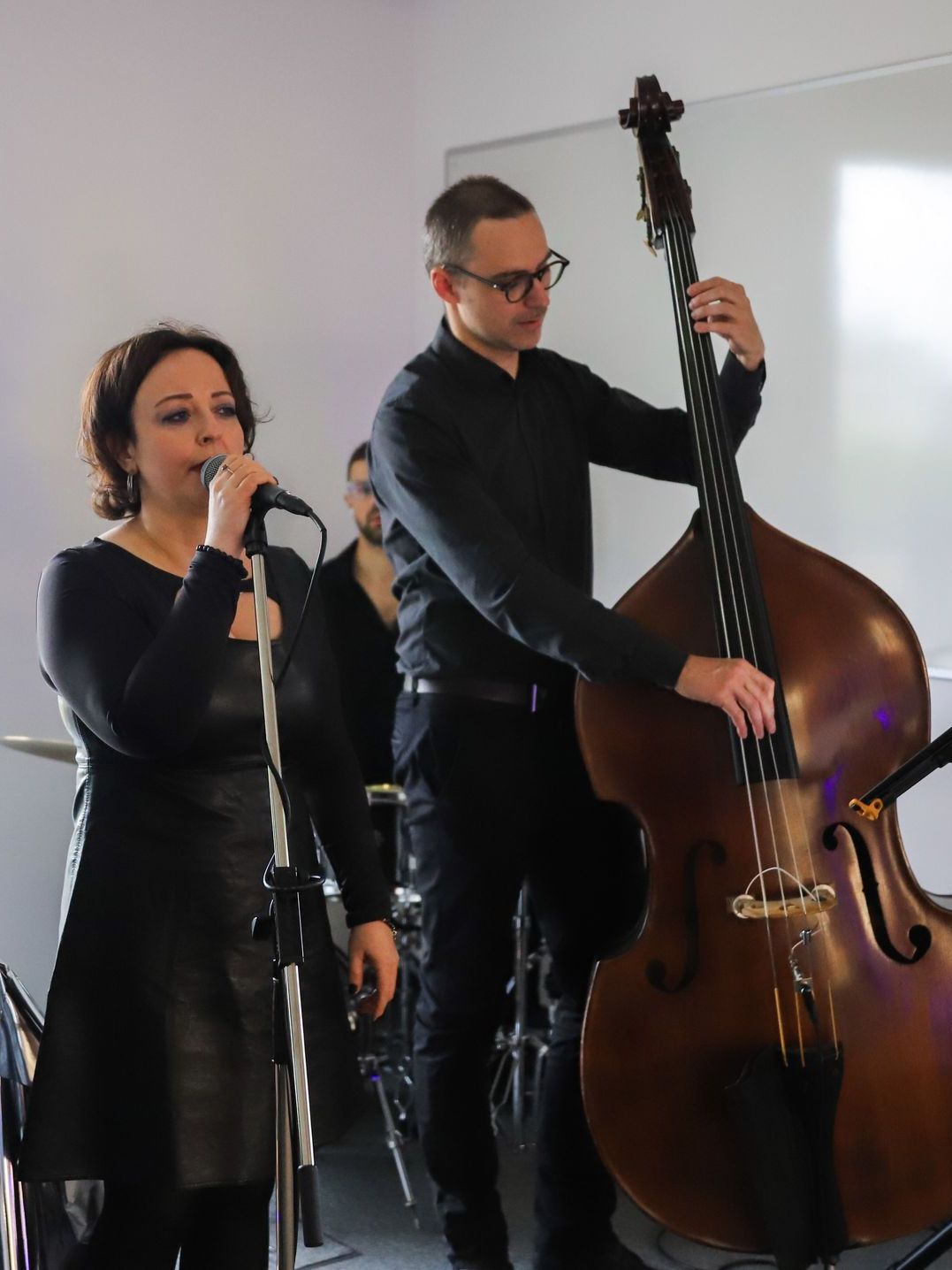A band performs. A woman sings into a microphone, a man plays a double bass, and a drummer is visible behind.