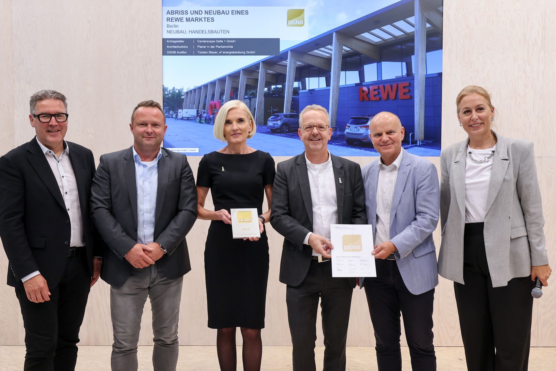 Sechs Personen posieren mit Auszeichnungen vor einer REWE-Filiale.
