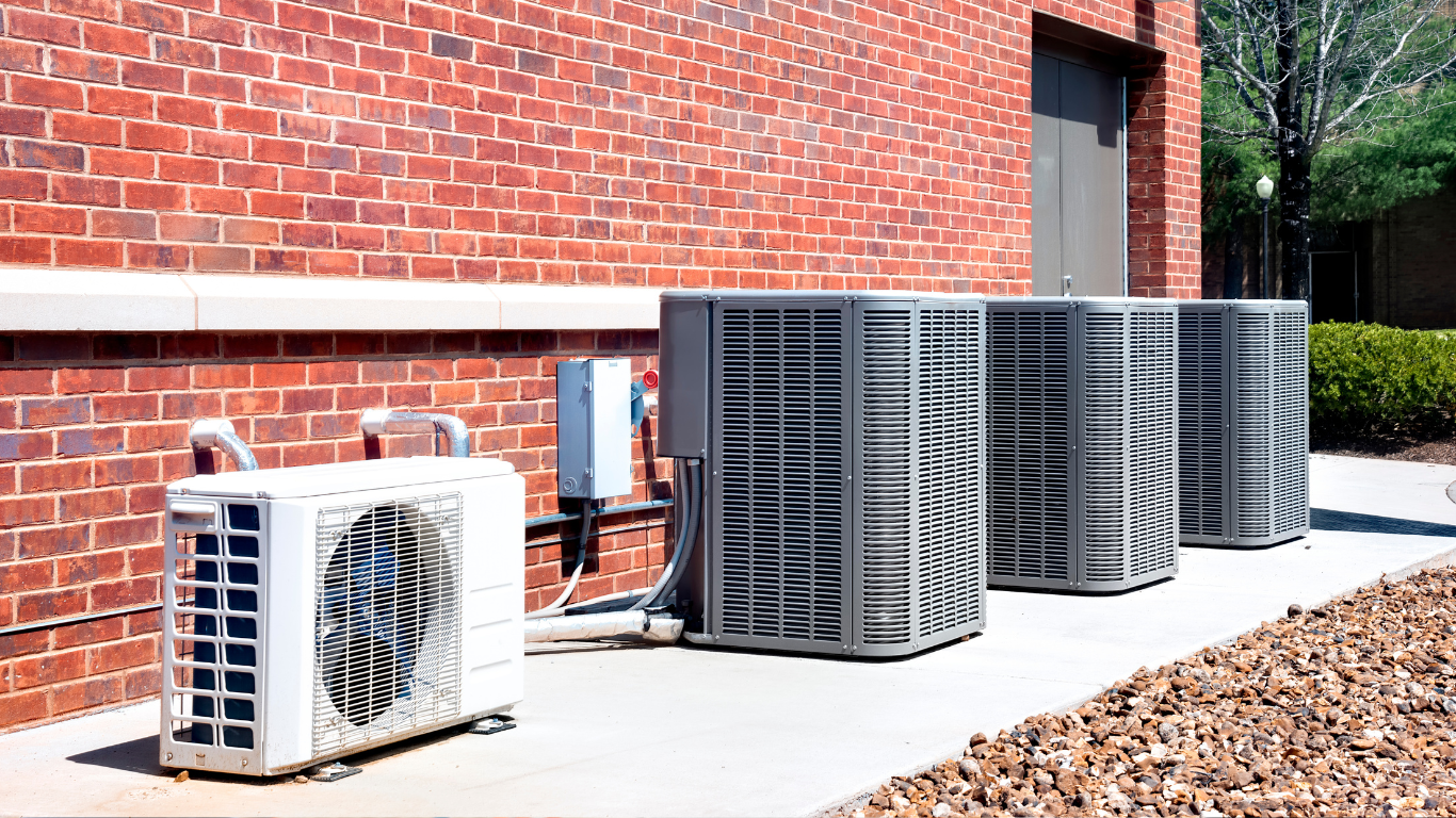 Multiple outdoor HVAC units beside a red brick building, with one smaller white unit in front.