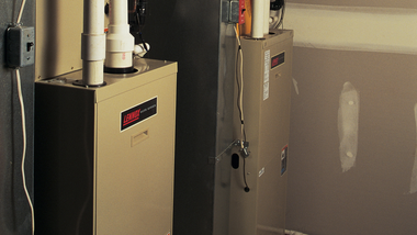 Two beige water heaters beside a wall with exposed pipes in a utility room