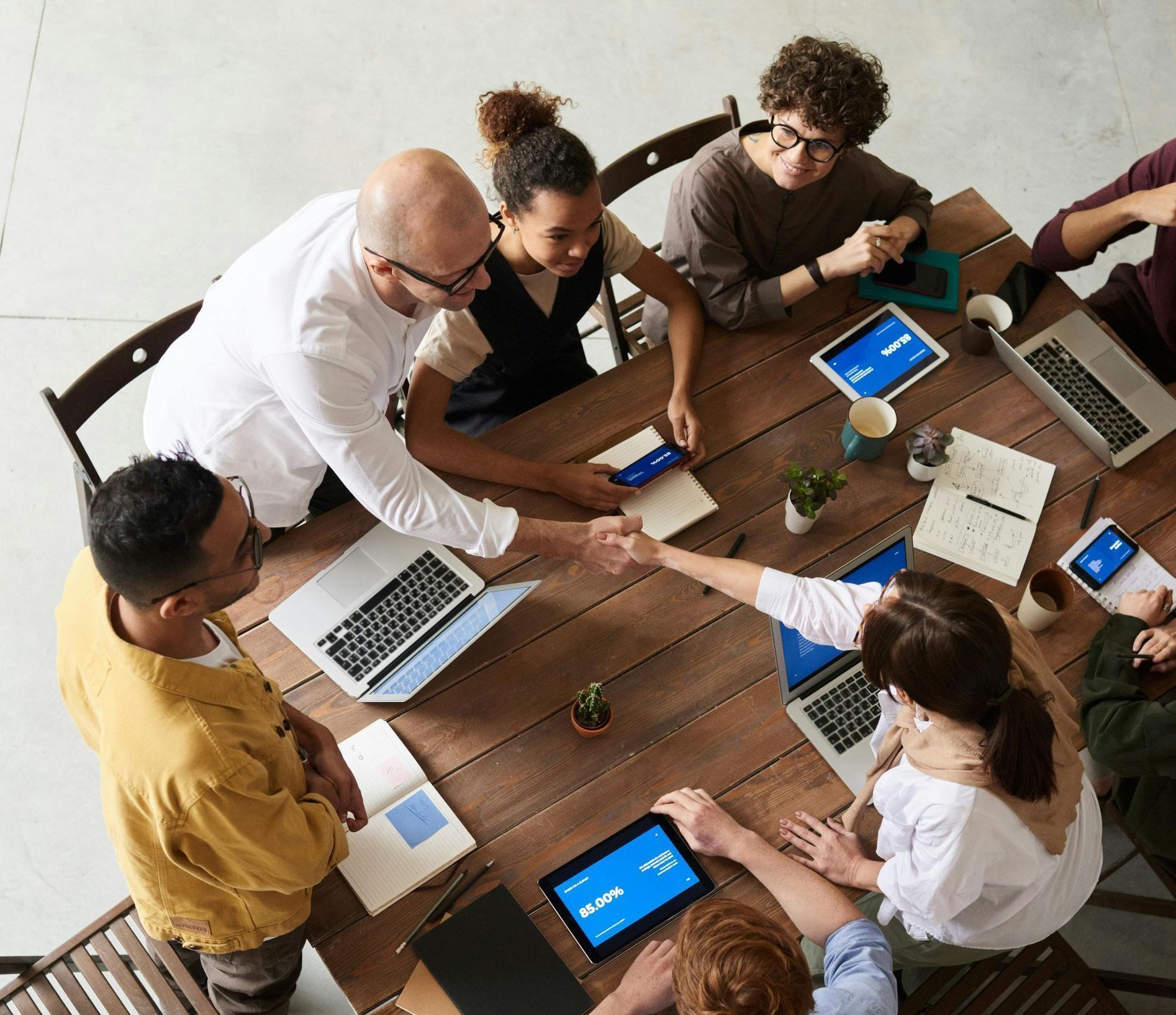 People shaking hands around a wooden table with laptops and tablets.