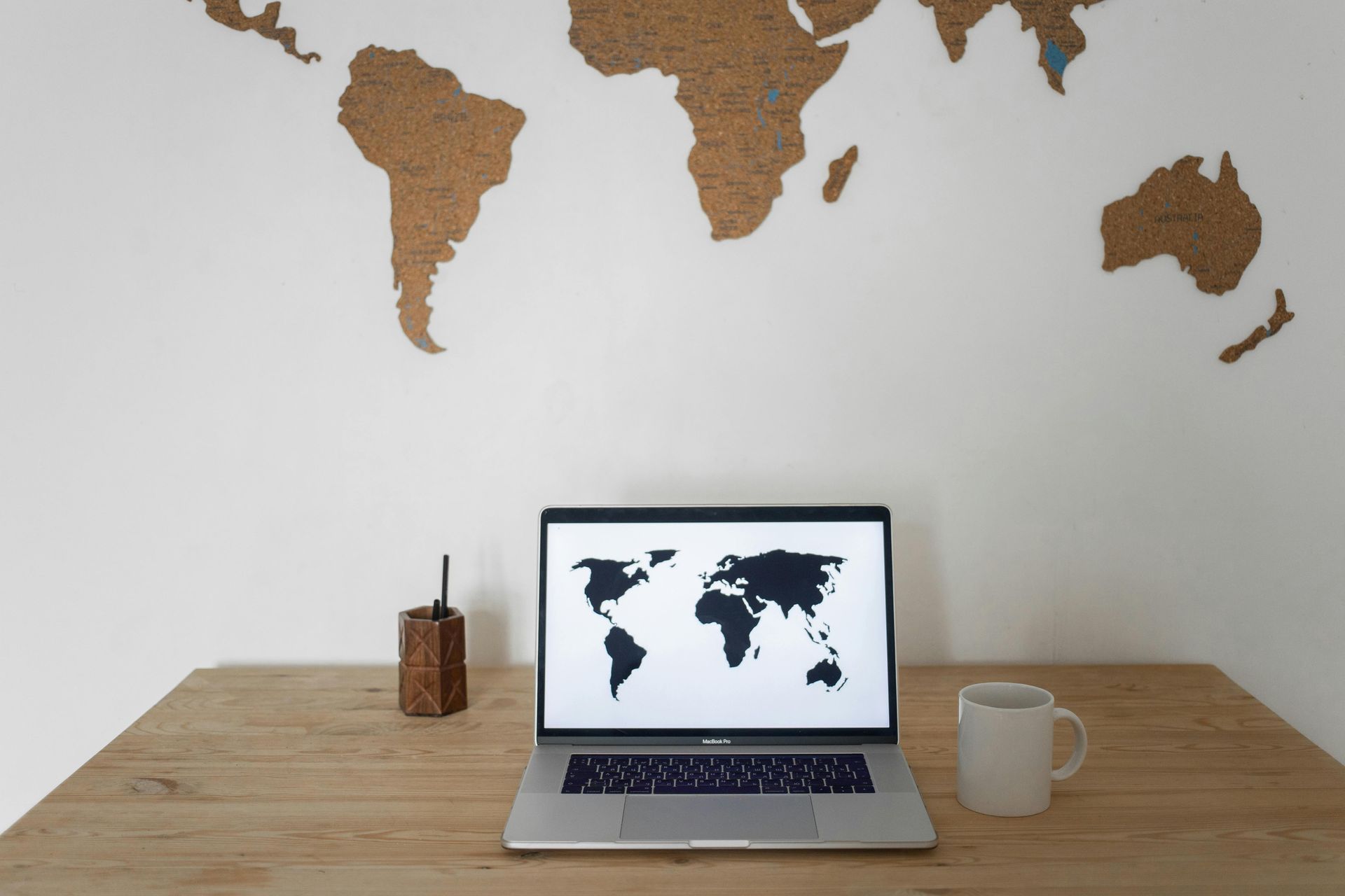 Laptop on a wooden desk displays world map, with cork world map on white wall behind.