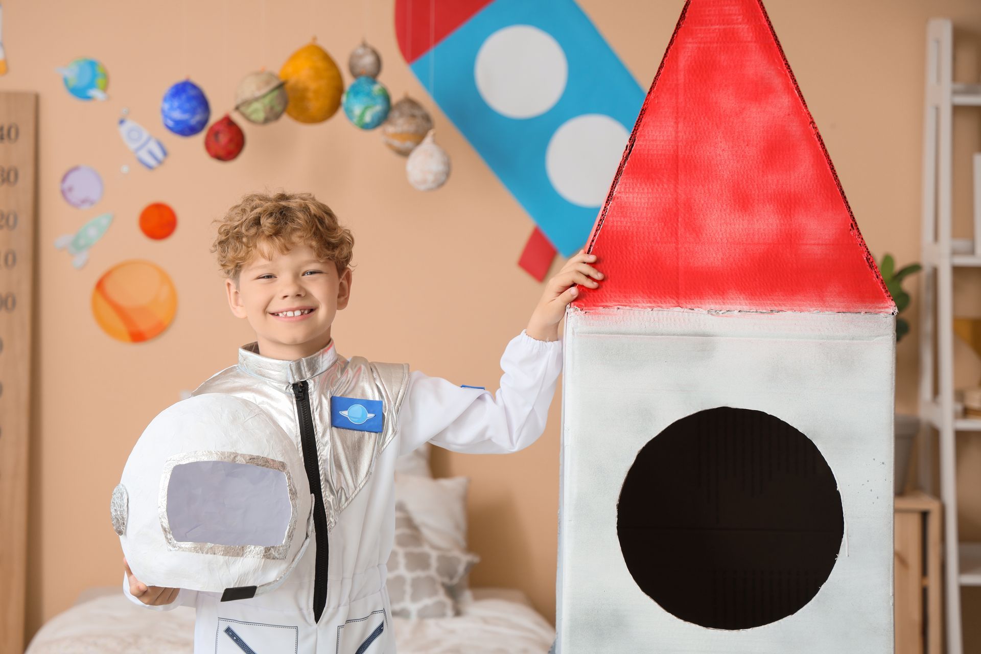 Child in a spacesuit smiling beside a cardboard rocket with a red tip in a decorated room