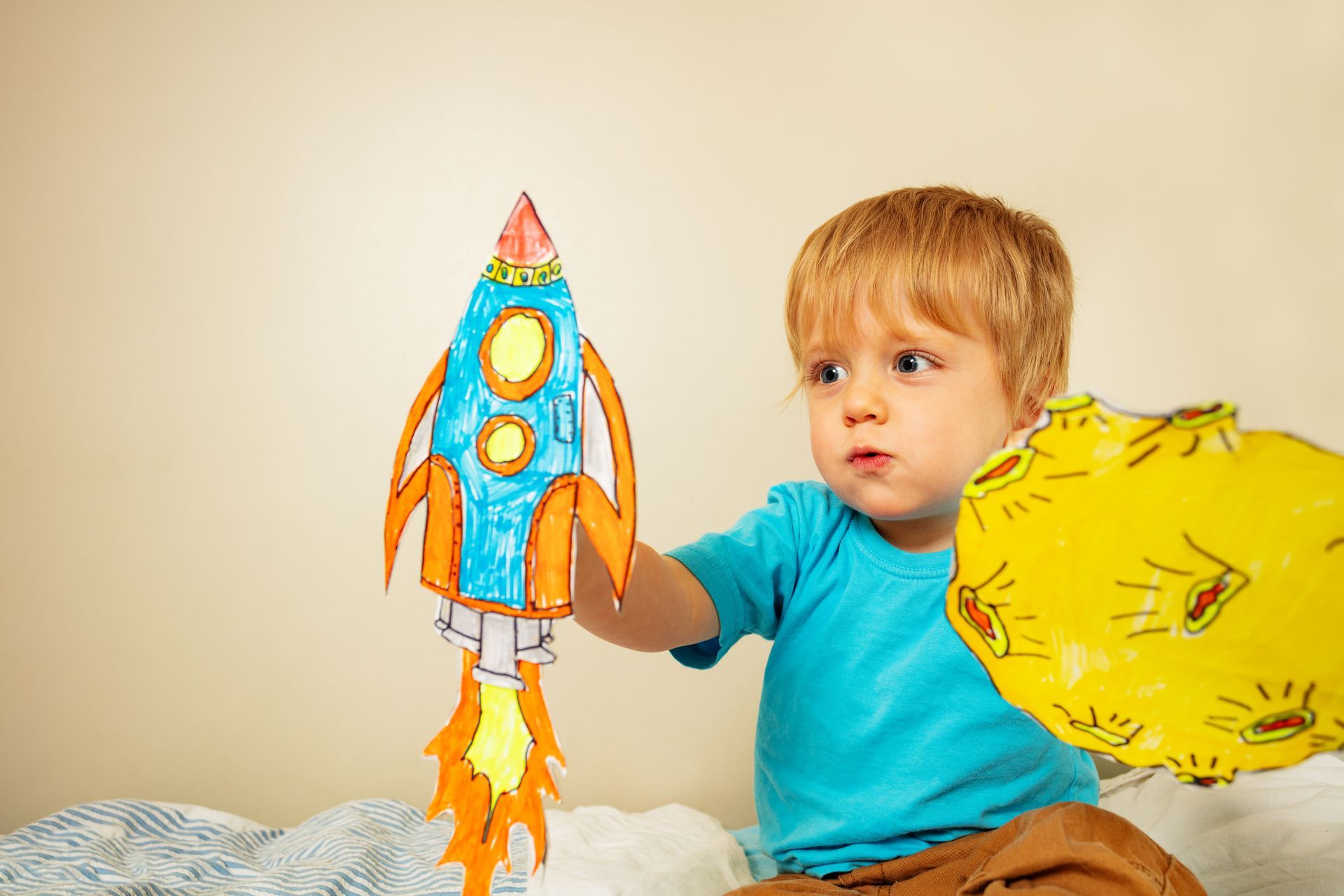 Child holding a colorful paper rocket and yellow cutout, seated indoors on a bed