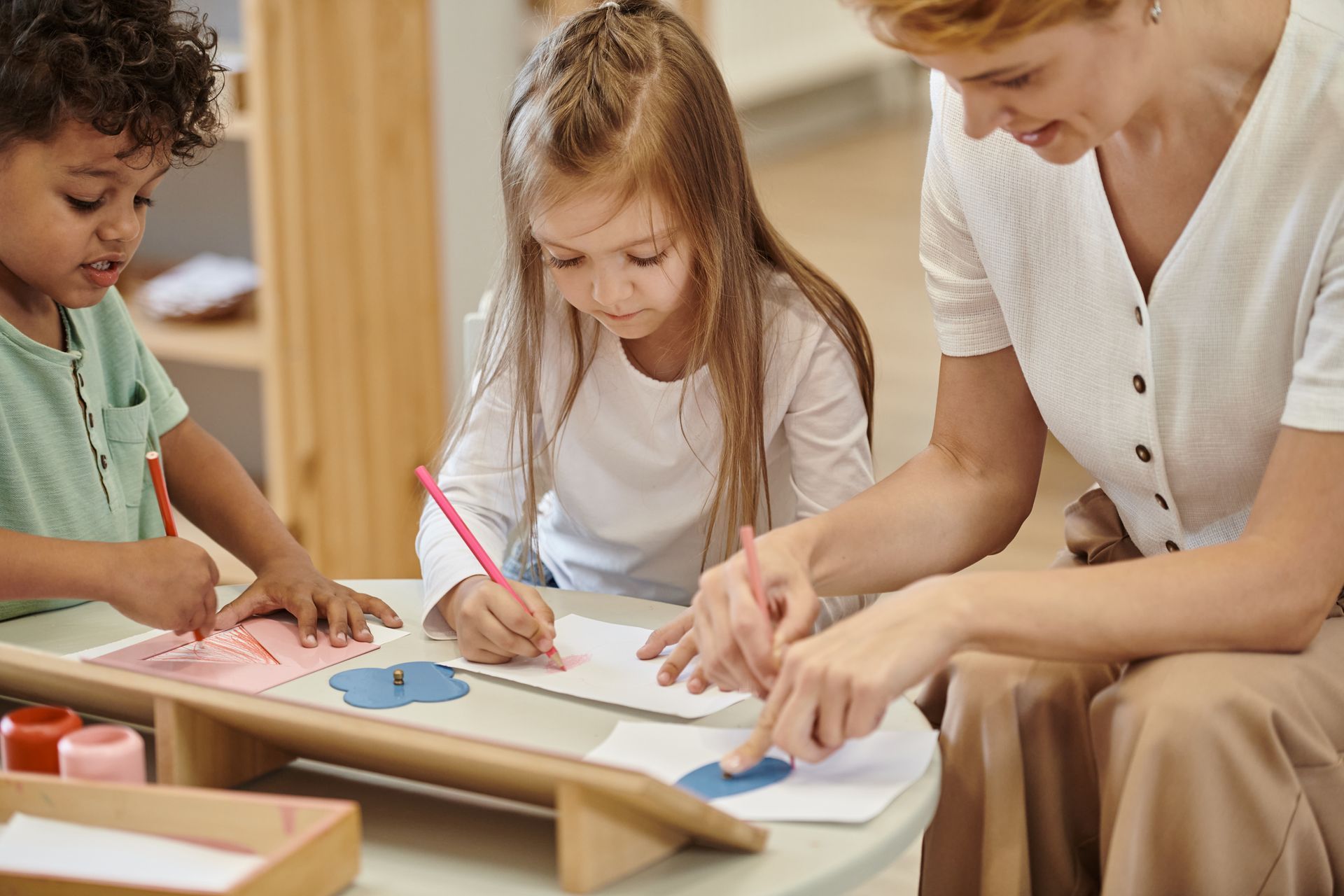 Two kids are drawing on a paper while a teacher watches them. Two kids are drawing on a paper while a teacher watches them.