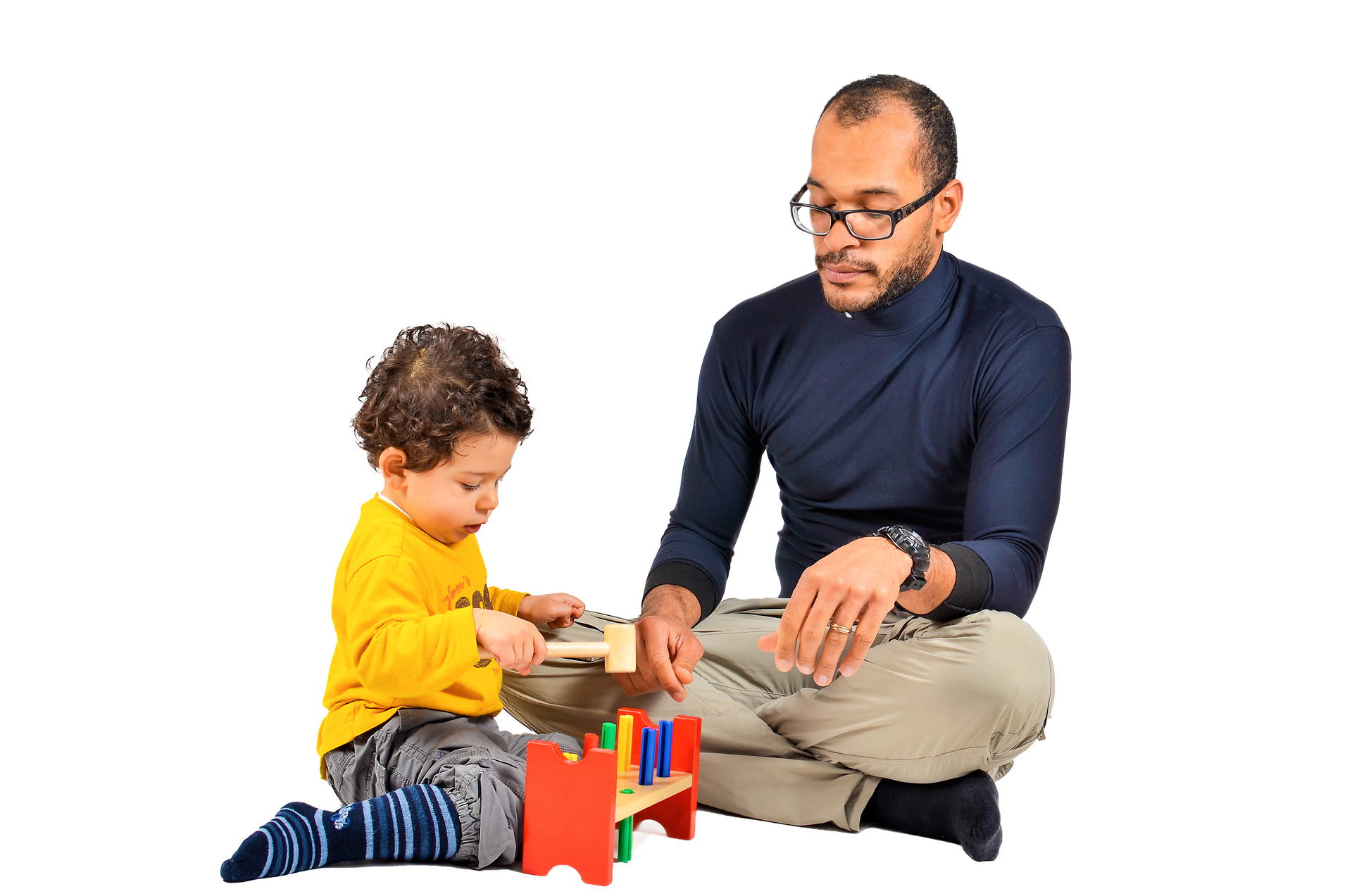 Man and young child playing with wooden toy. Man watches child hammering pegs. A man with glasses watches a child play with a mallet and colorful wooden toy, both seated on the floor.