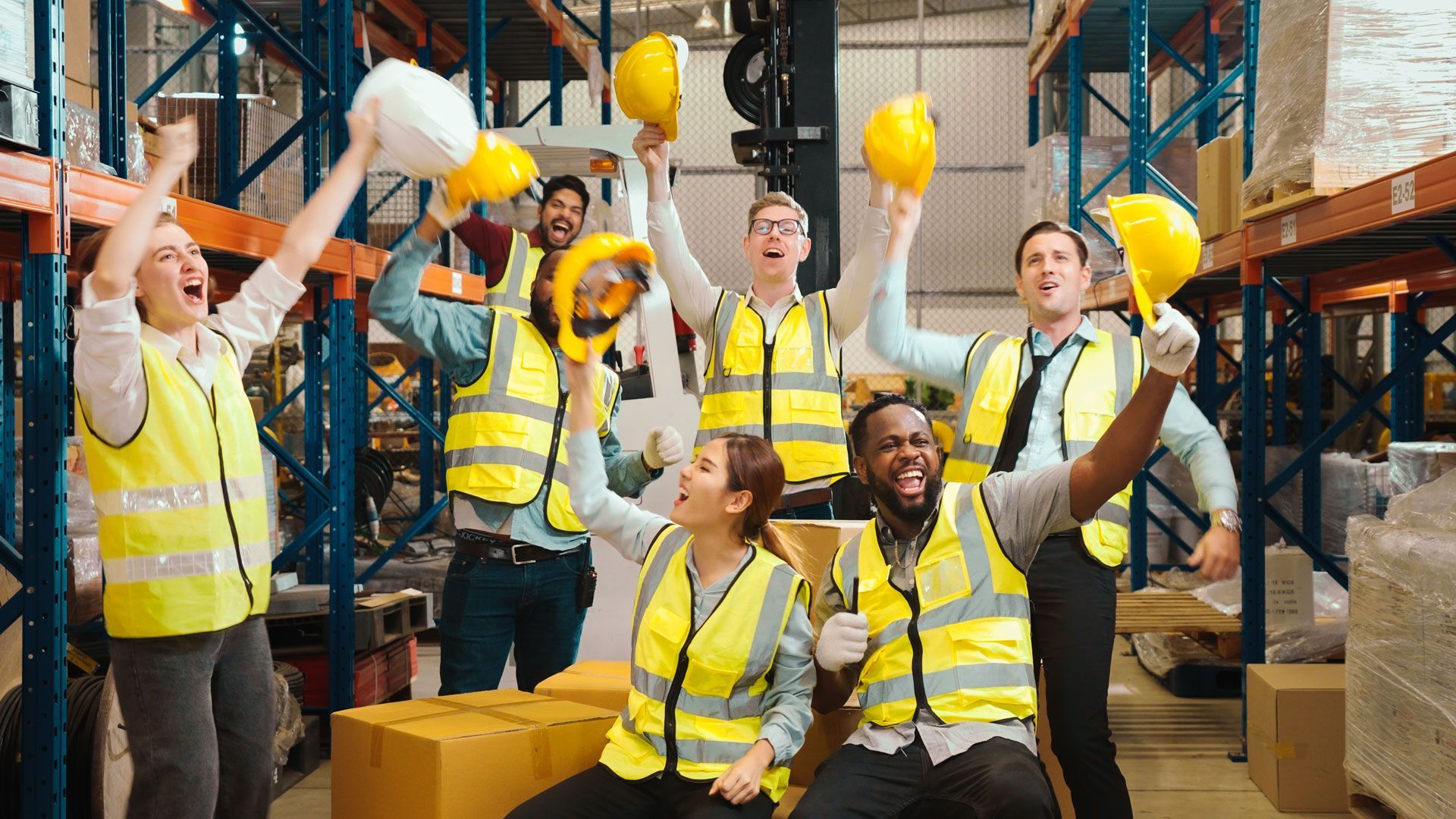 Warehouse workers in yellow safety vests celebrating with raised hard hats among stacked boxes