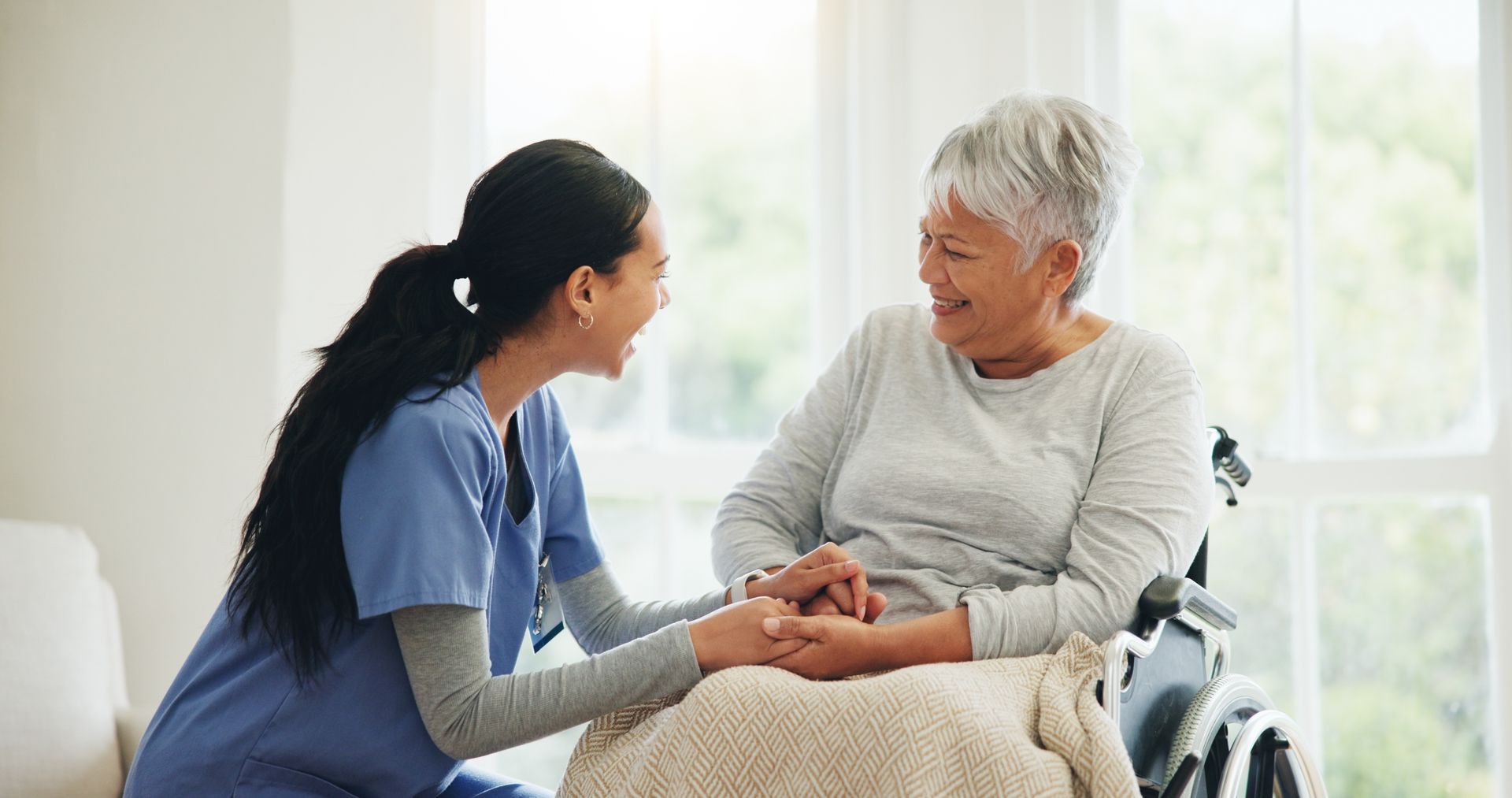 A caregiver in blue scrubs holds hands with a smiling person in a wheelchair in a bright room.