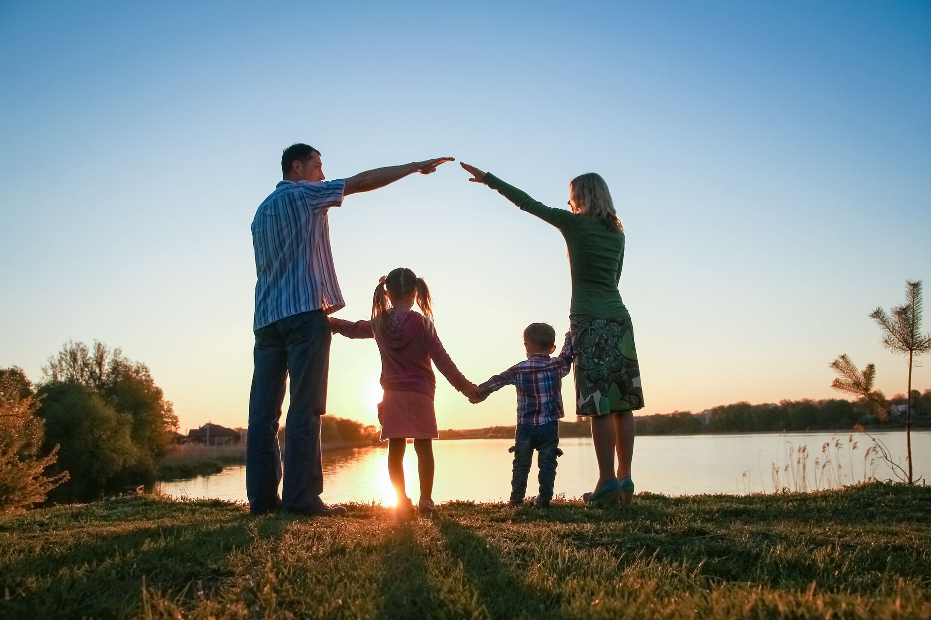 A family stands by a lake at sunset, forming the shape of a house roof with their joined hands overhead.