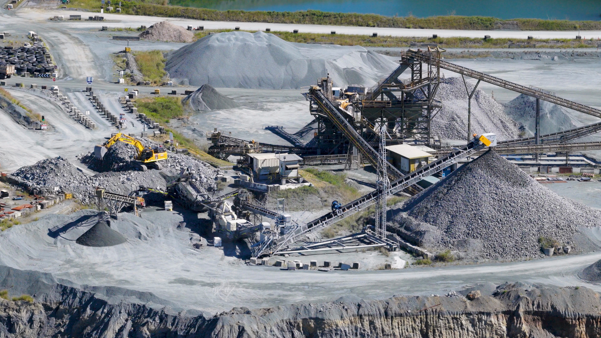 Open-pit mining quarry with conveyors, gravel piles, and heavy machinery under a clear sky