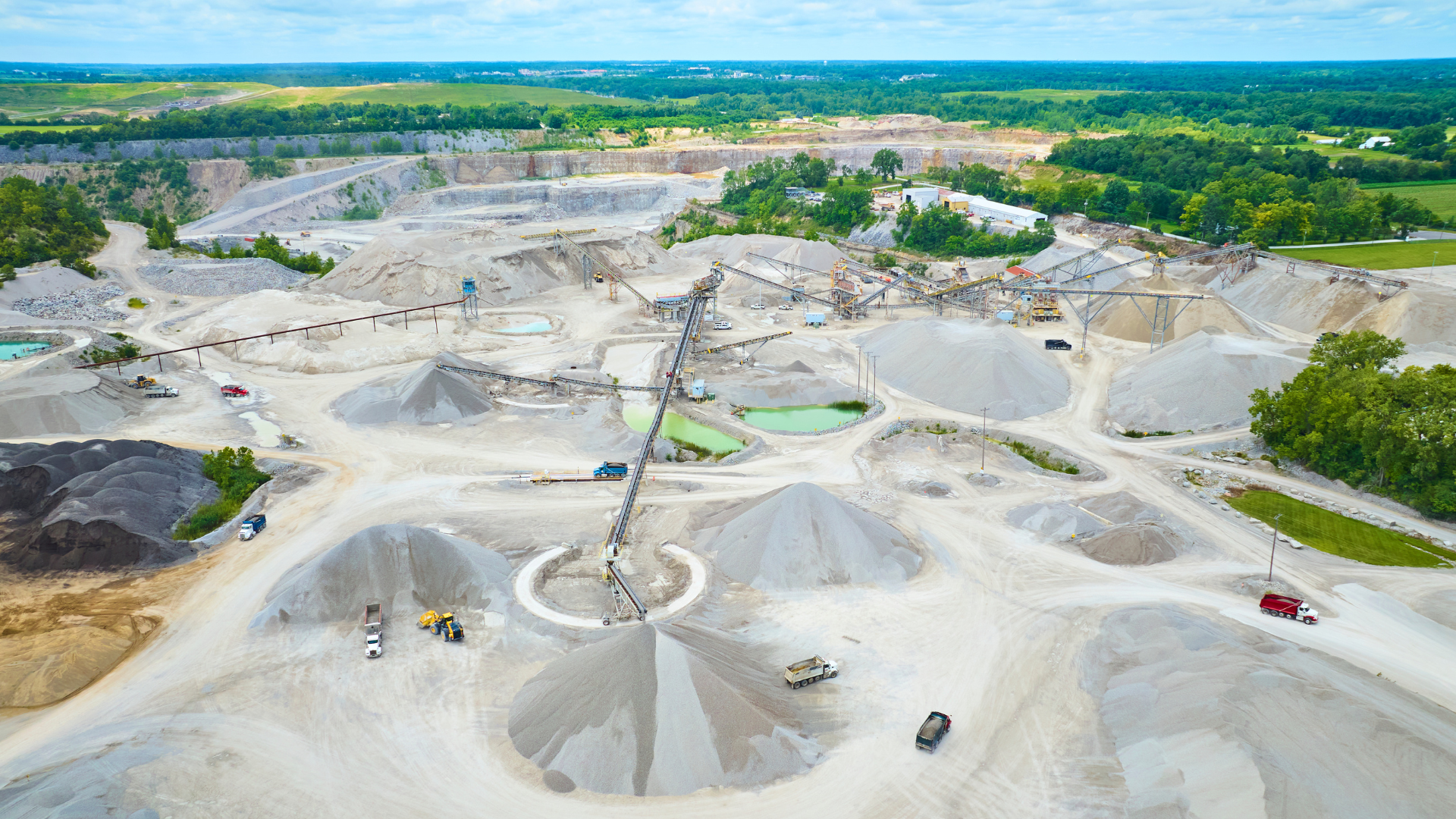 Aerial view of a large gray quarry with trucks, sand piles, and green forest beyond.