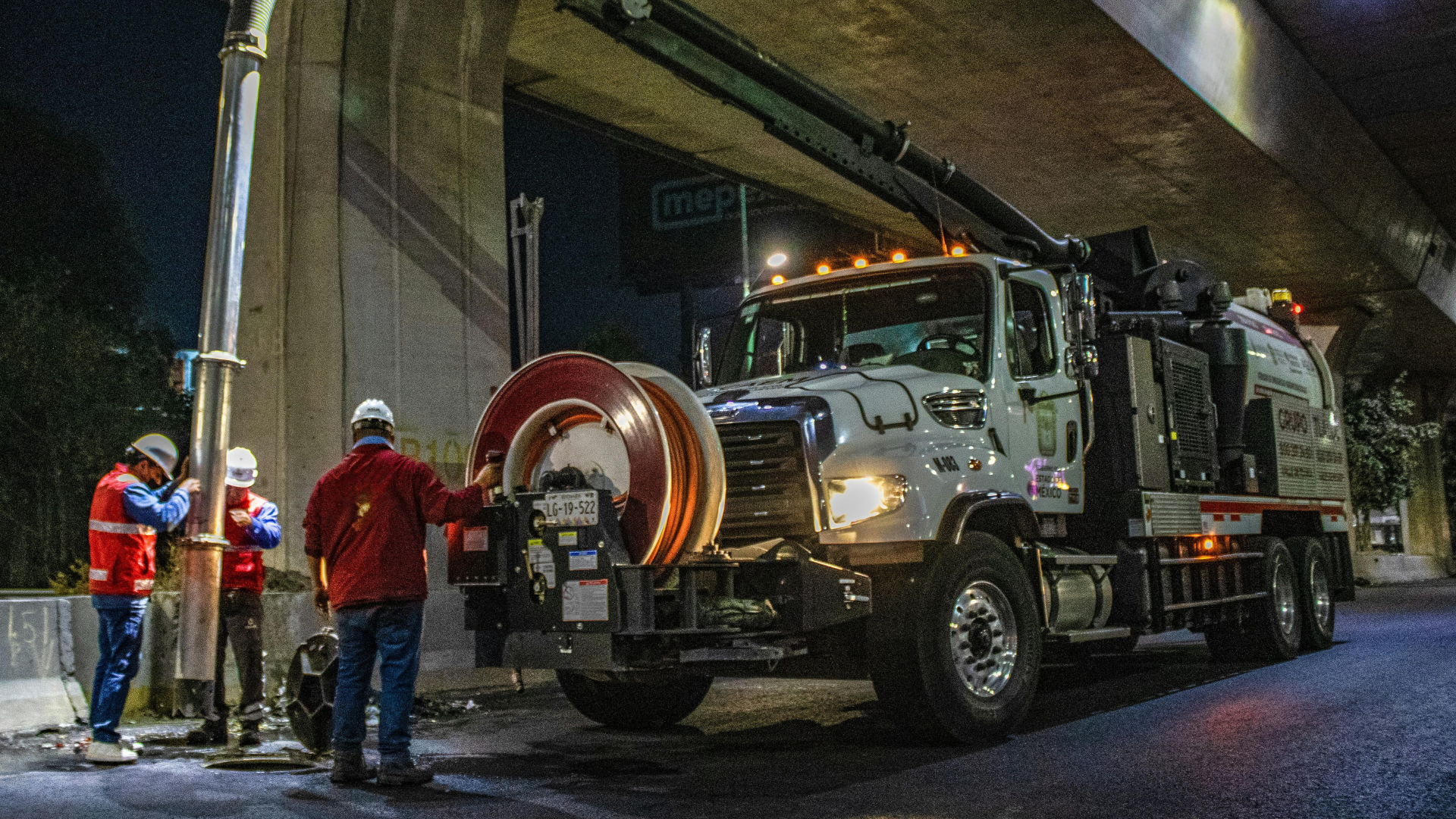 Utility truck and workers in hard hats under an overpass at night, with traffic cones and equipment.