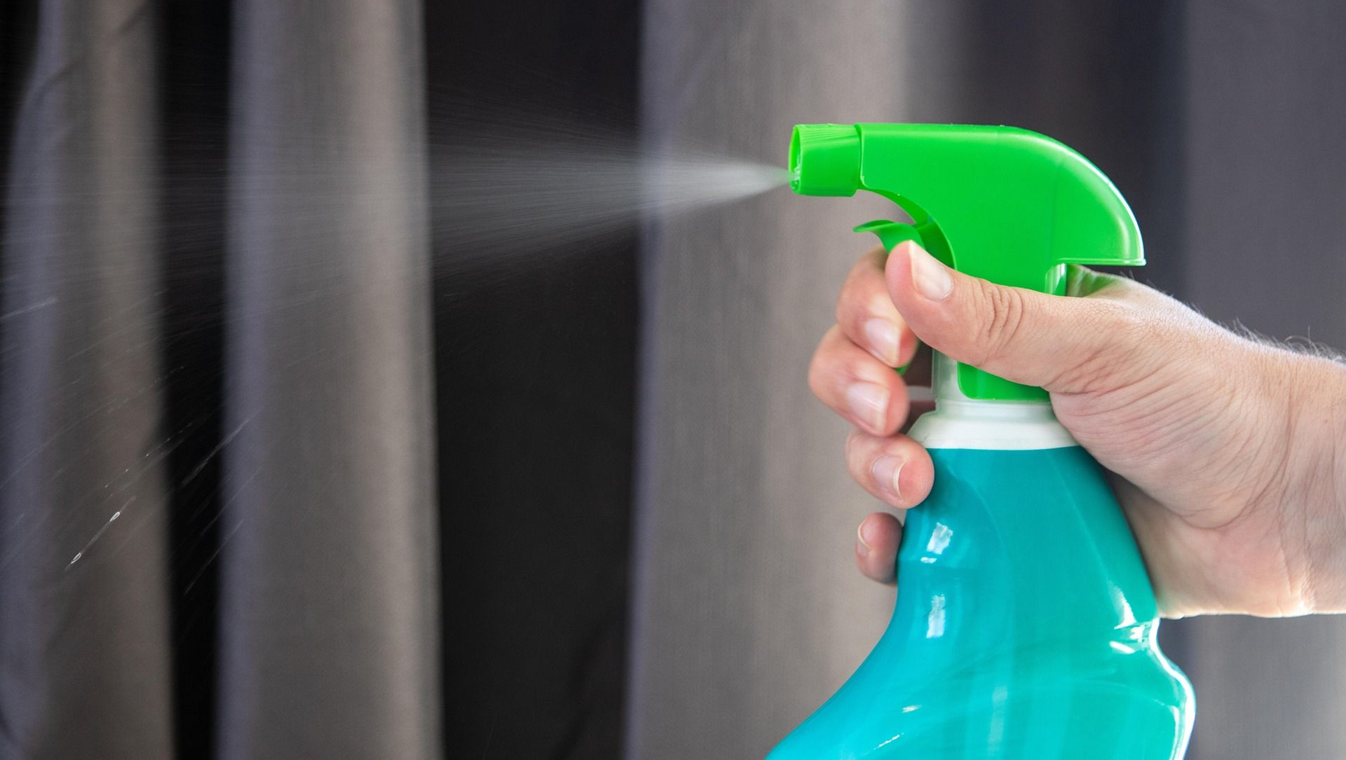 Hand spraying a green and blue bottle towards dark gray striped curtains.