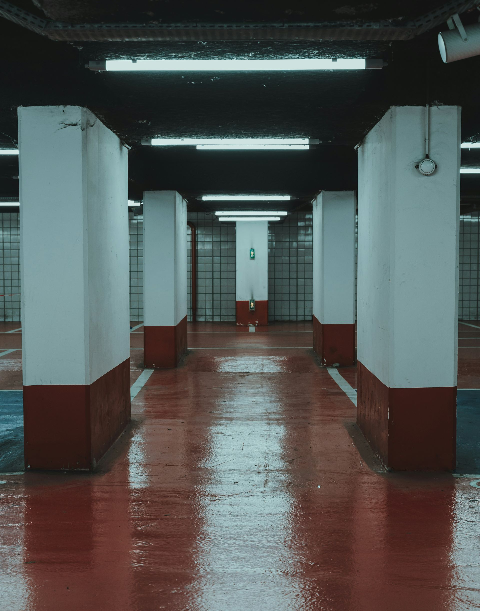 Empty parking garage, red floor reflecting lights, white pillars with red bases, somber atmosphere.