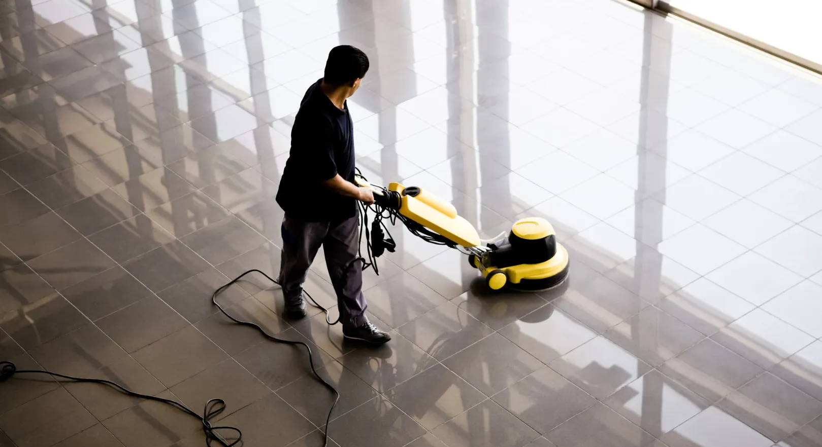 Person using a yellow floor polisher on a glossy gray floor near a window.