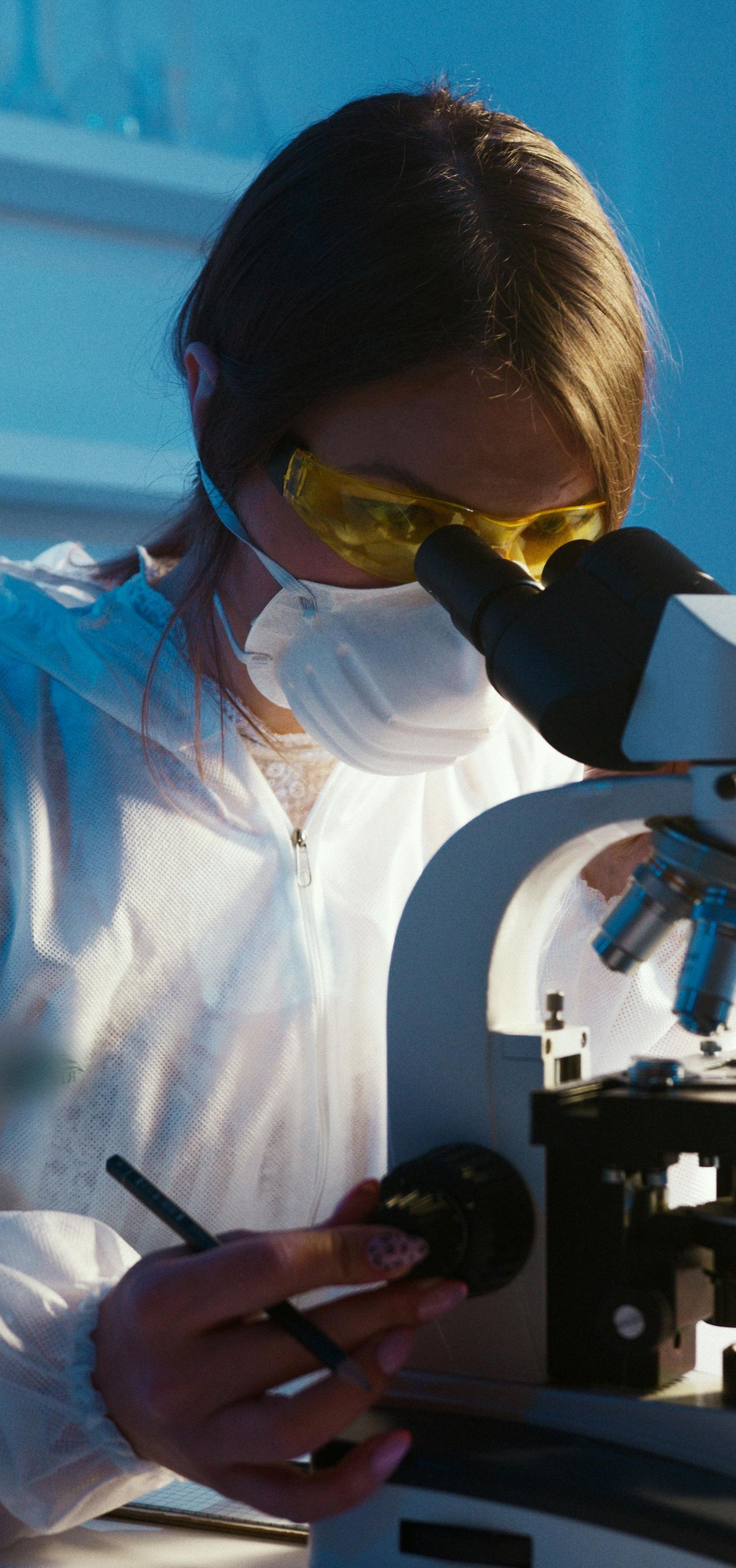 Person in protective gear looking through a microscope, holding a dark circular object and pen.