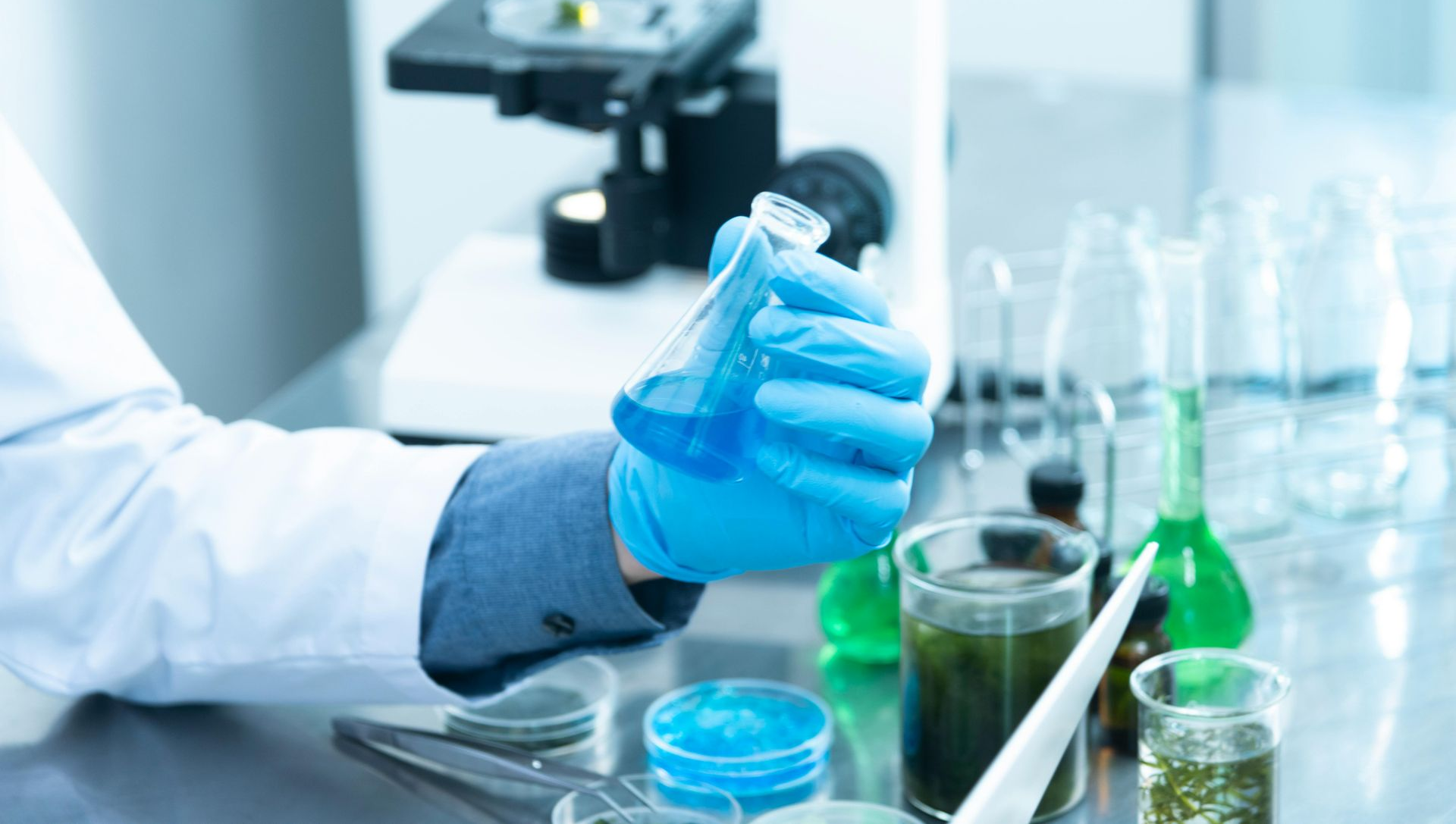 Scientist wearing gloves holding blue liquid in a flask, lab setting with microscope and glassware.
