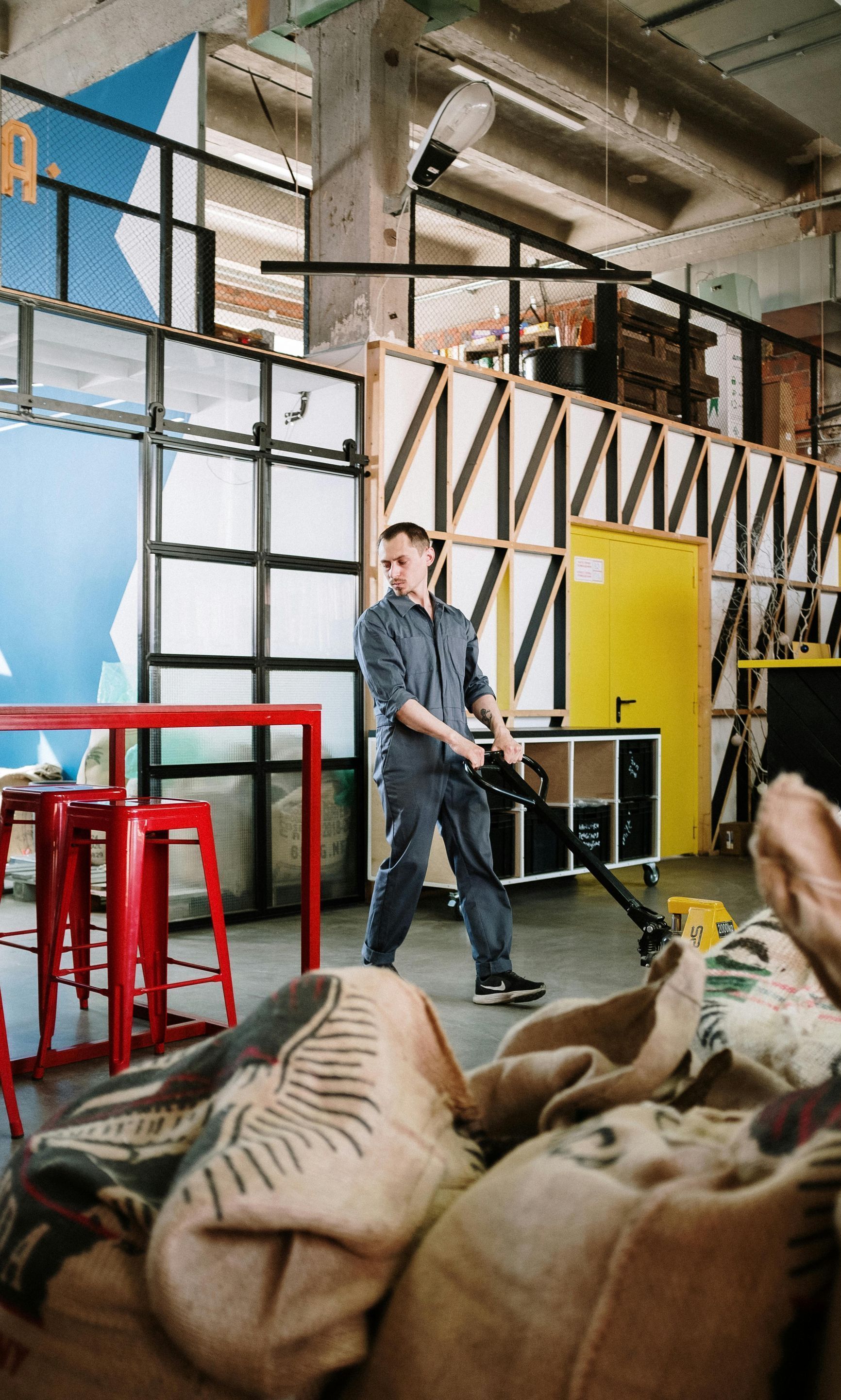 Person in blue jumpsuit holding a rifle, walking through an industrial-style space with burlap sacks in the foreground.