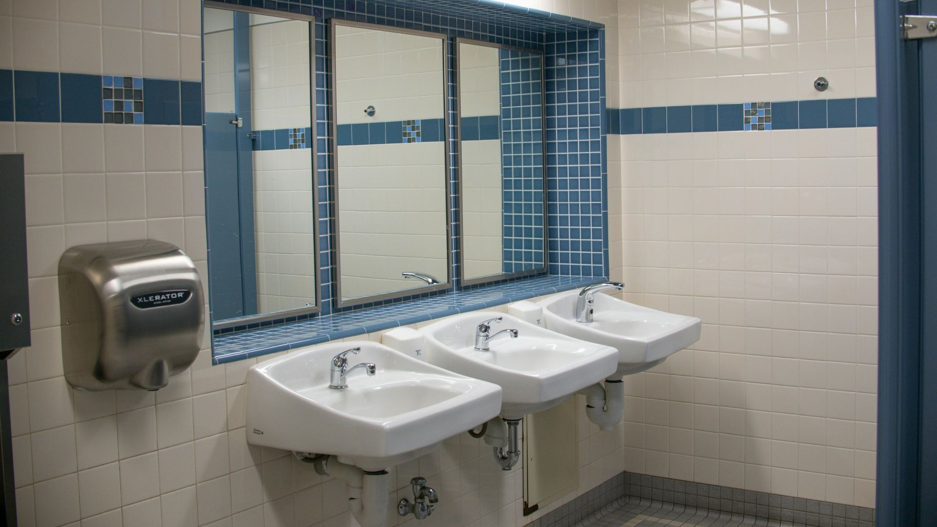 Public restroom with three sinks, mirrors, and a hand dryer, white and blue tiled walls.