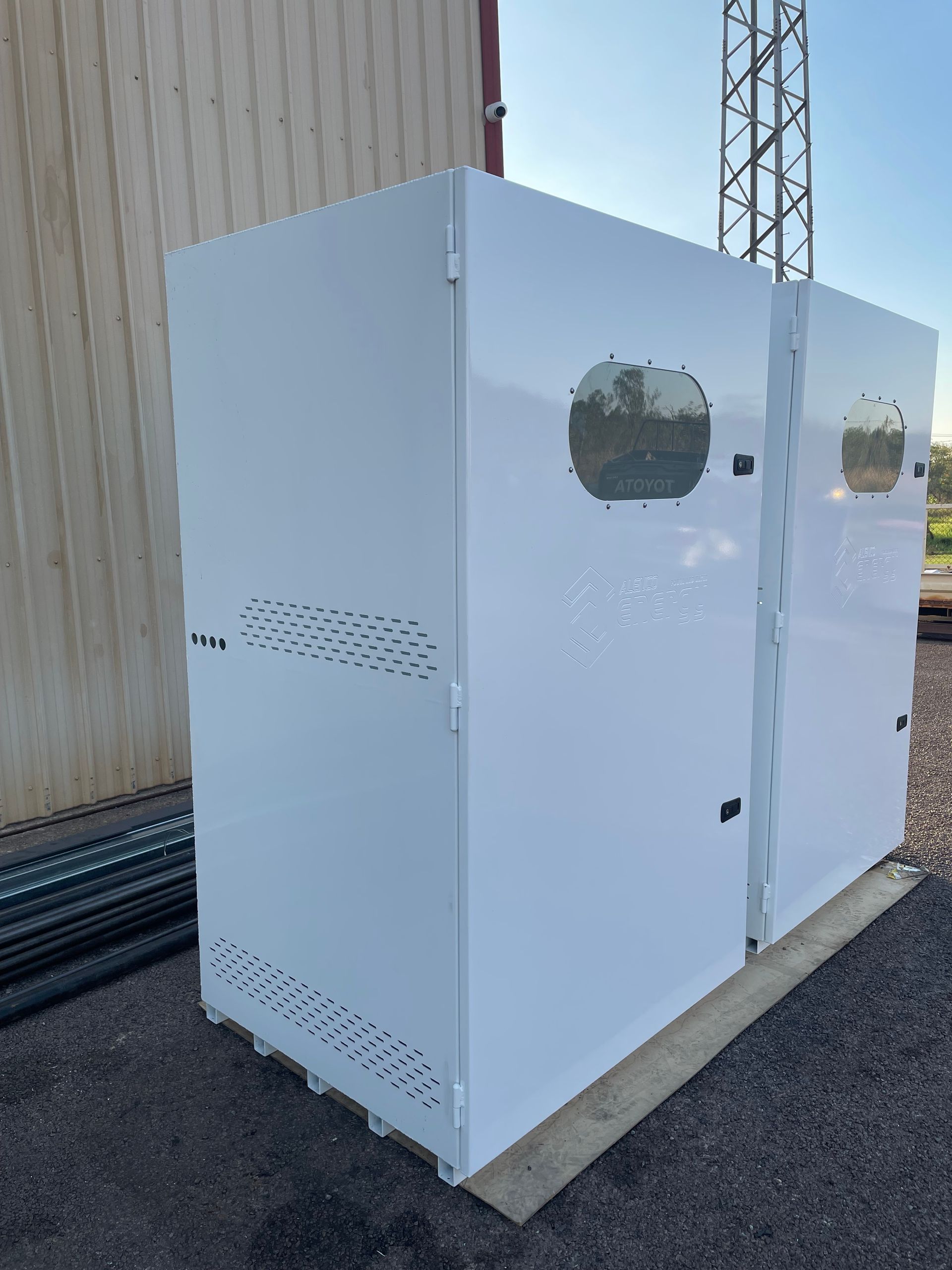 White metal enclosure with small windows beside a beige industrial building outdoors.