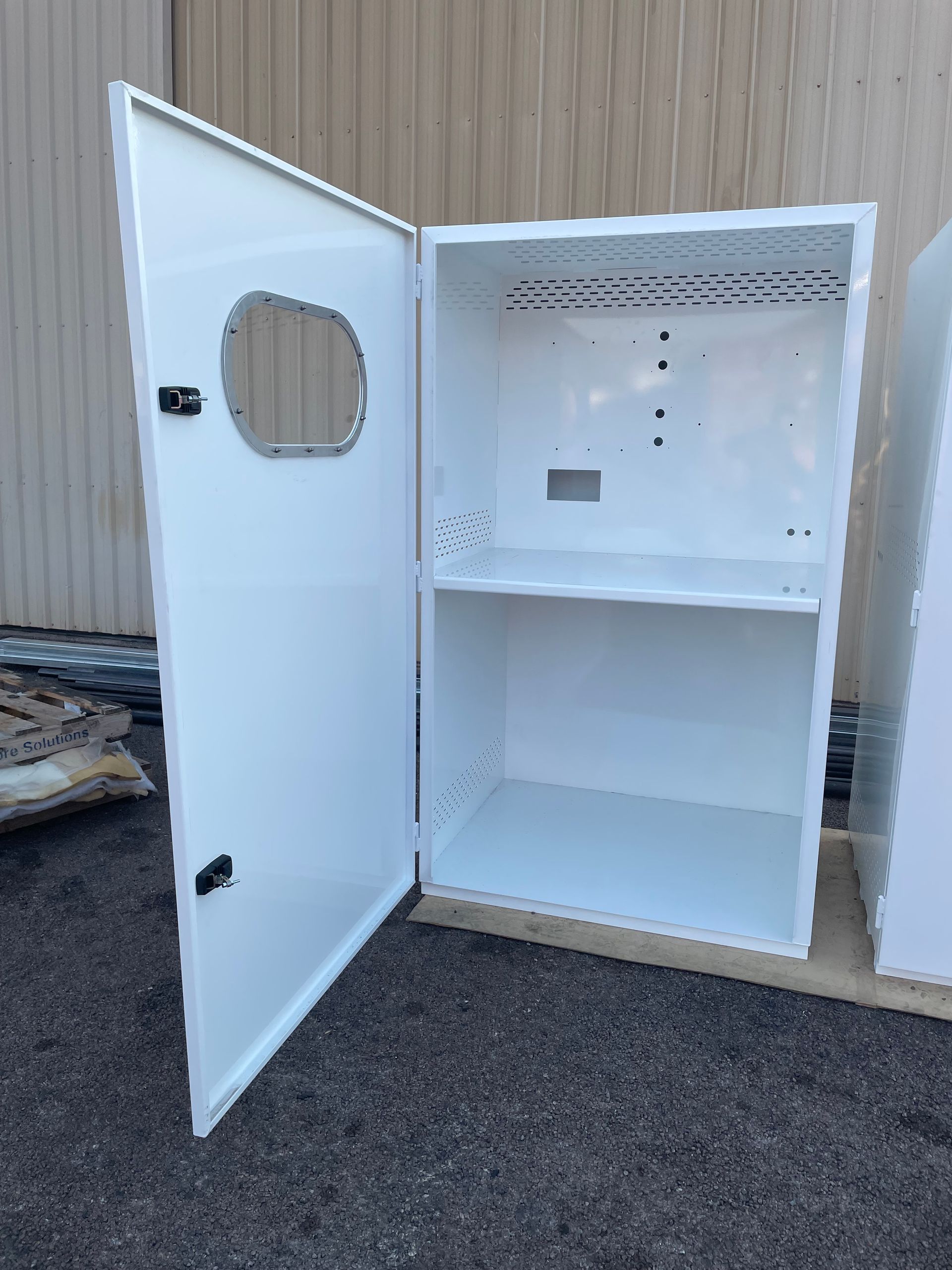 White metal cabinet with open door, pegboard back, and shelf, standing outdoors on pavement.
