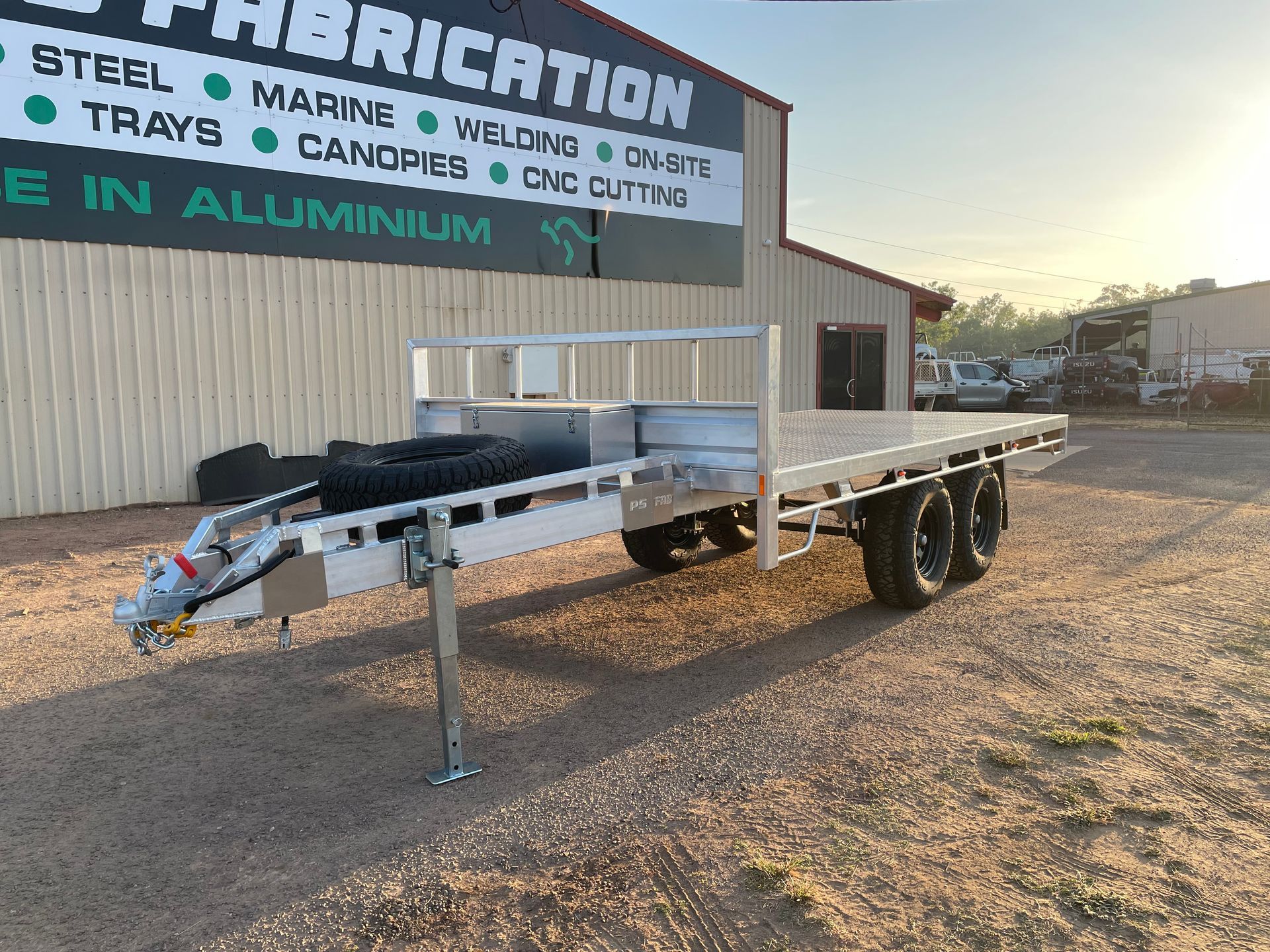 Empty aluminum utility trailer parked on gravel beside a fabrication shop at sunset
