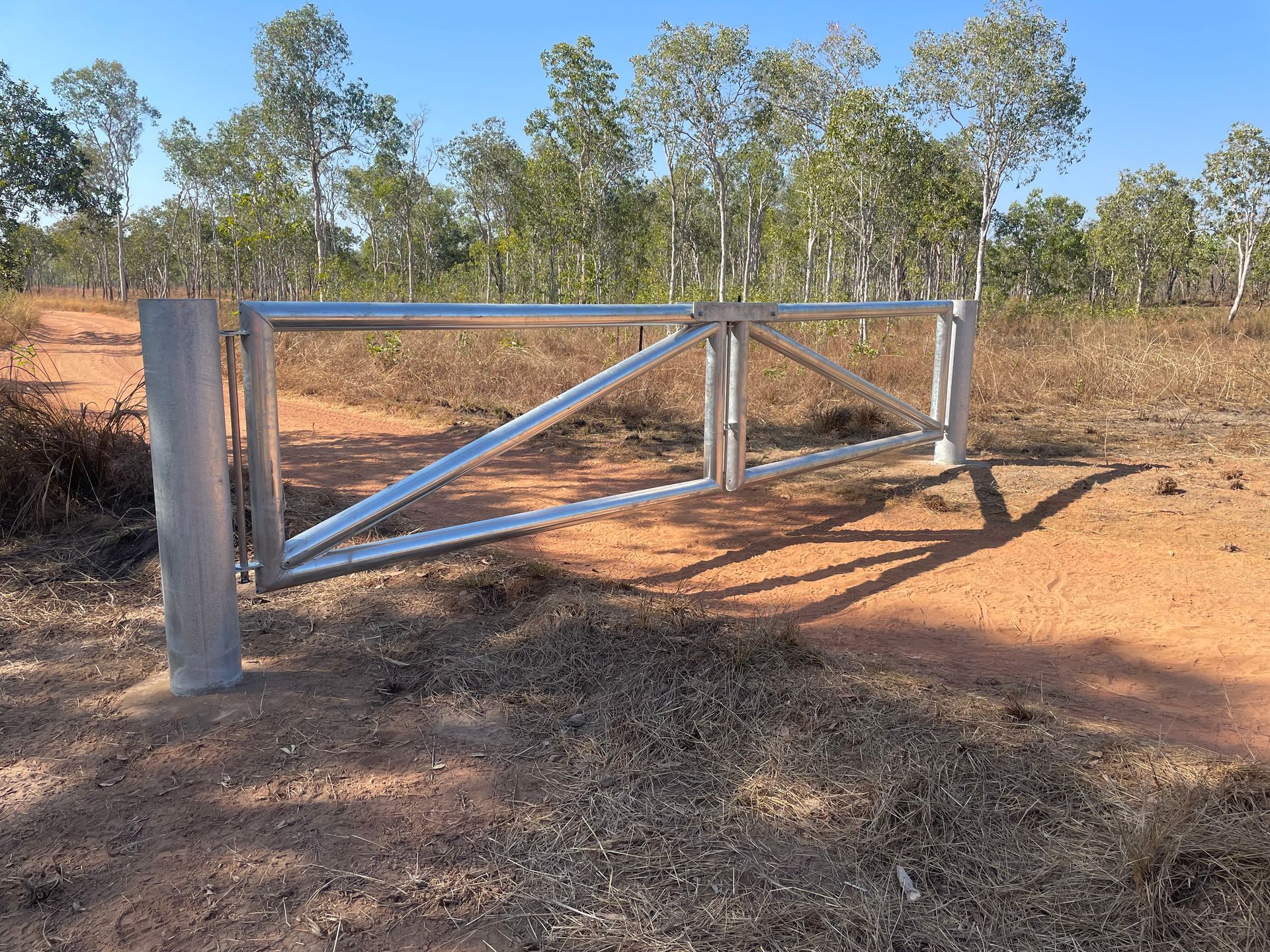 Metal farm gate with diagonal brace on a dirt track, surrounded by dry grass and trees under a clear sky
