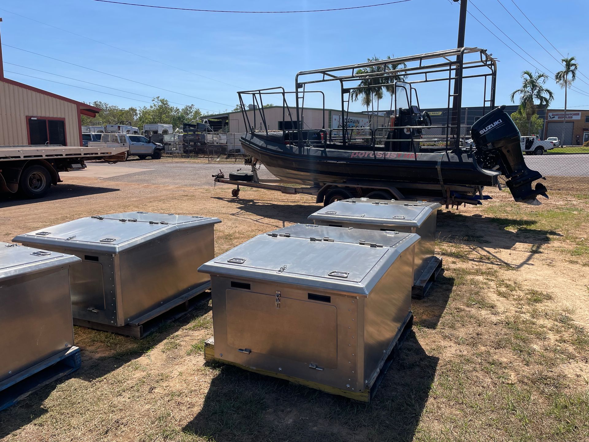 Two metal utility boxes in a dusty lot with a black boat on a trailer in the background