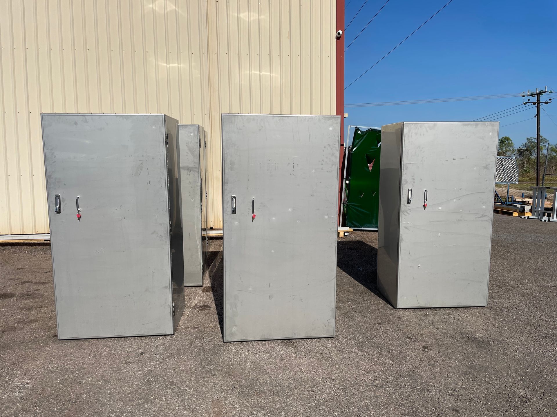 Three gray metal cabinets lined up outside a beige warehouse on a gravel lot under a blue sky