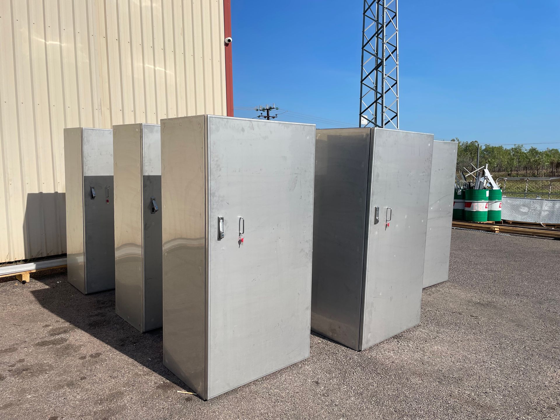 Gray utility cabinets outdoors beside a beige building, with blue sky and industrial equipment nearby
