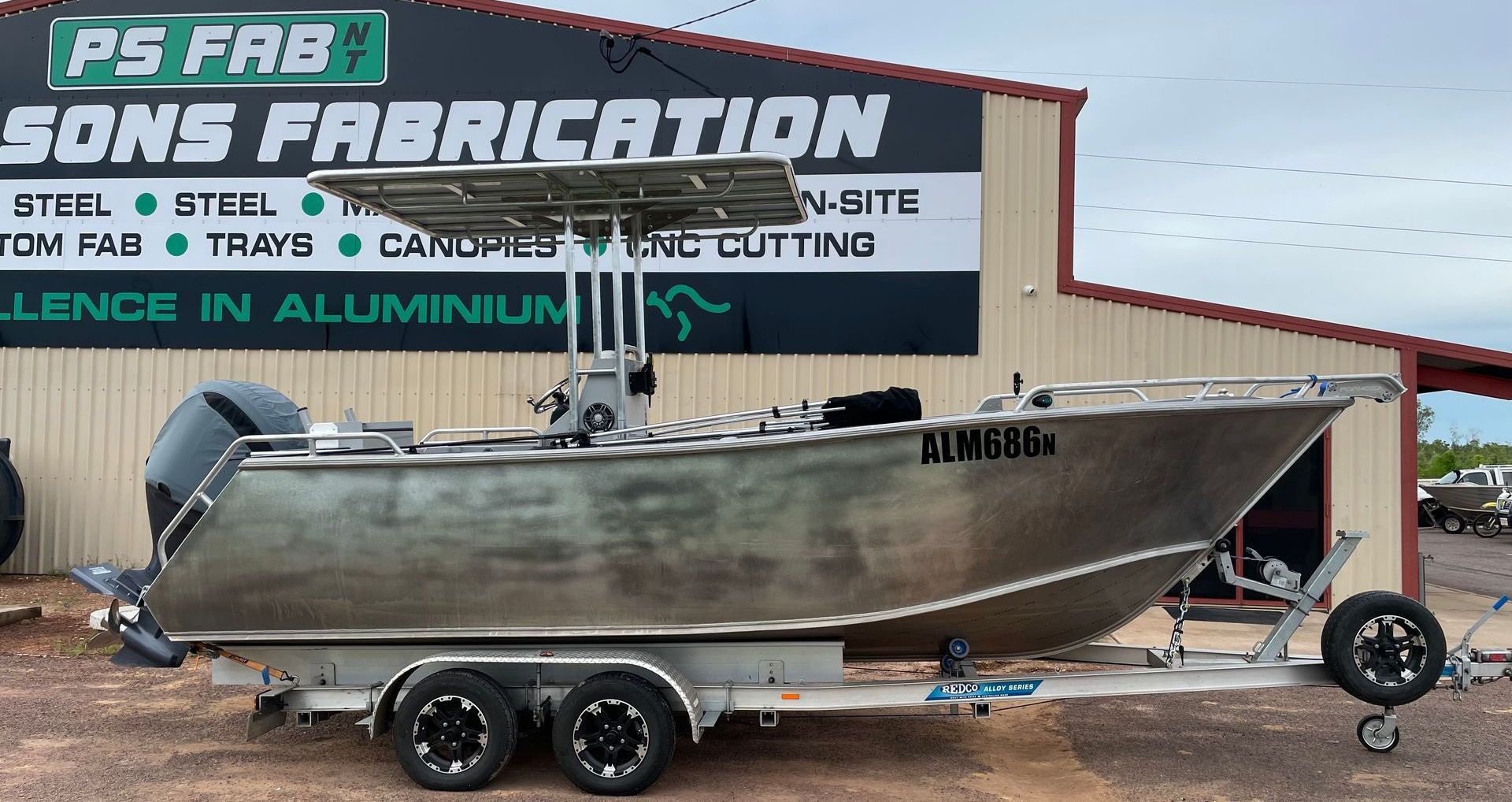 Aluminum boat on a trailer in front of a fabrication shop sign.