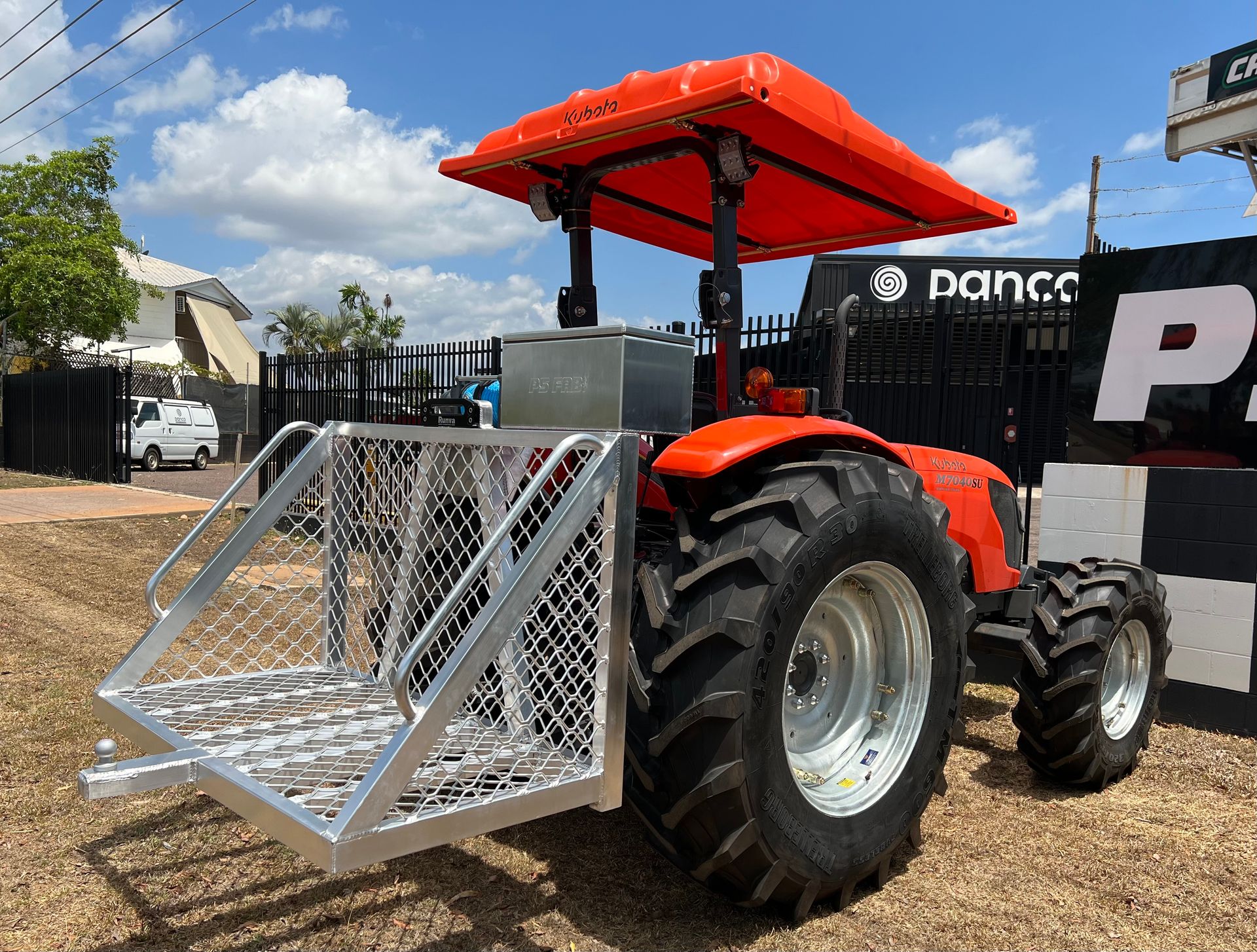 Red tractor with a front metal basket attachment parked outdoors on a dirt lot under a bright sky