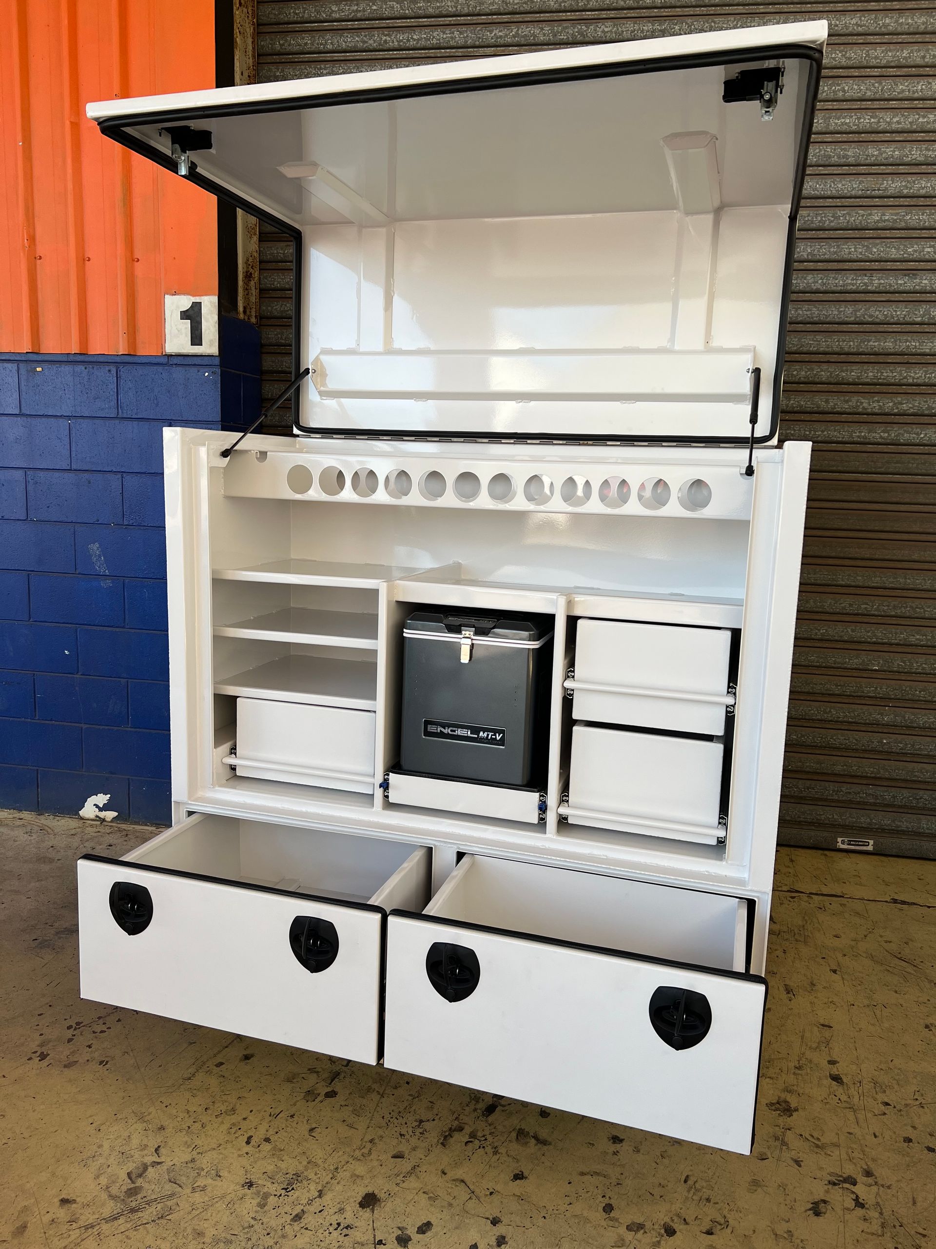 Open white kitchen cabinet with shelves, drawers, and a central appliance against a brick wall.