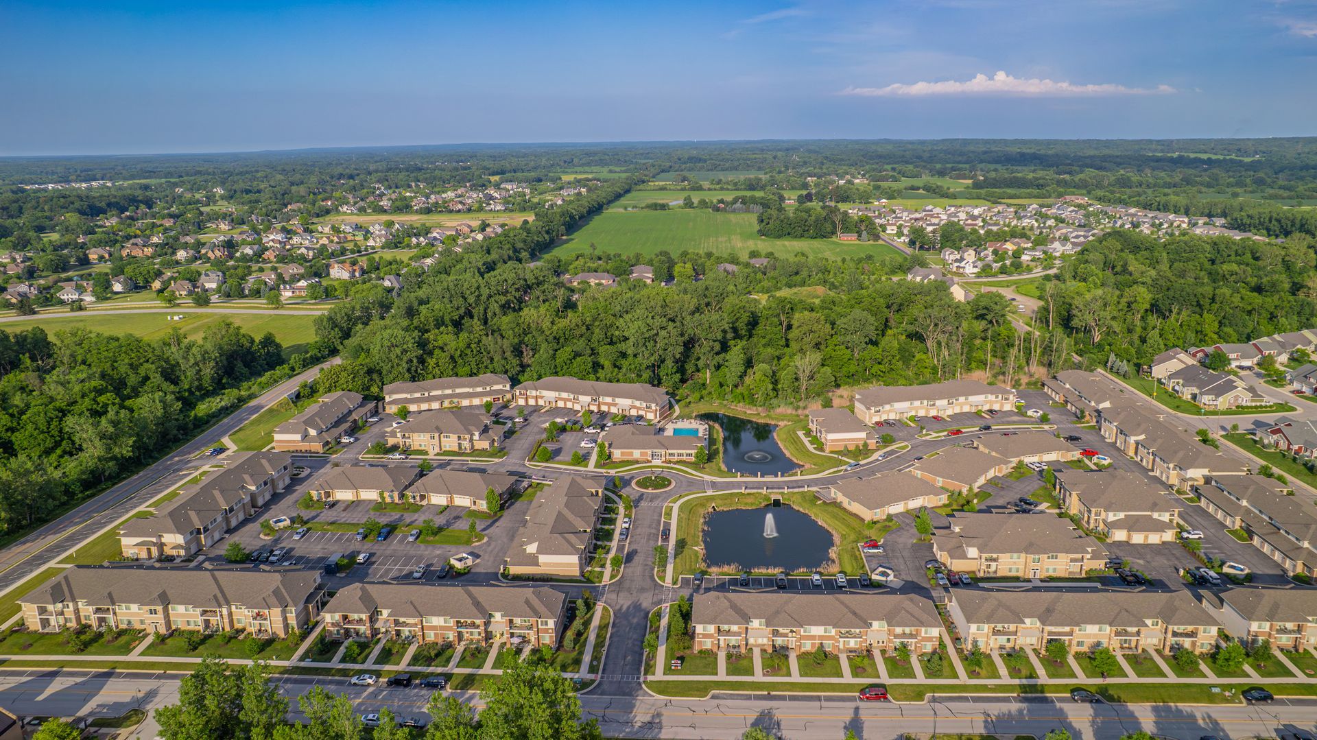 Aerial view of a suburban apartment complex, with multiple buildings, roads, and a pond surrounded by greenery.