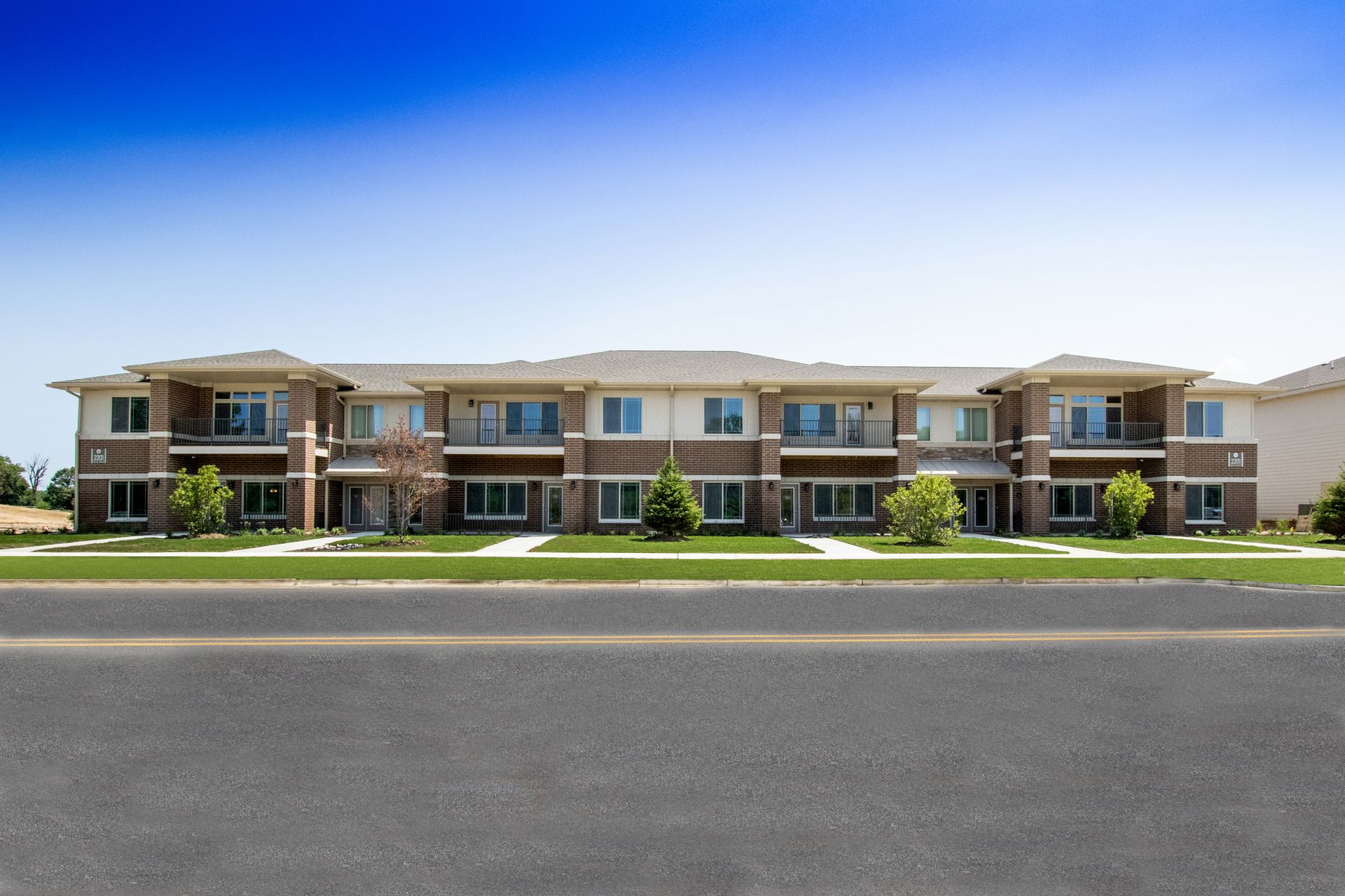 Two-story apartment building with tan siding, brown accents, and a blue sky in the background.