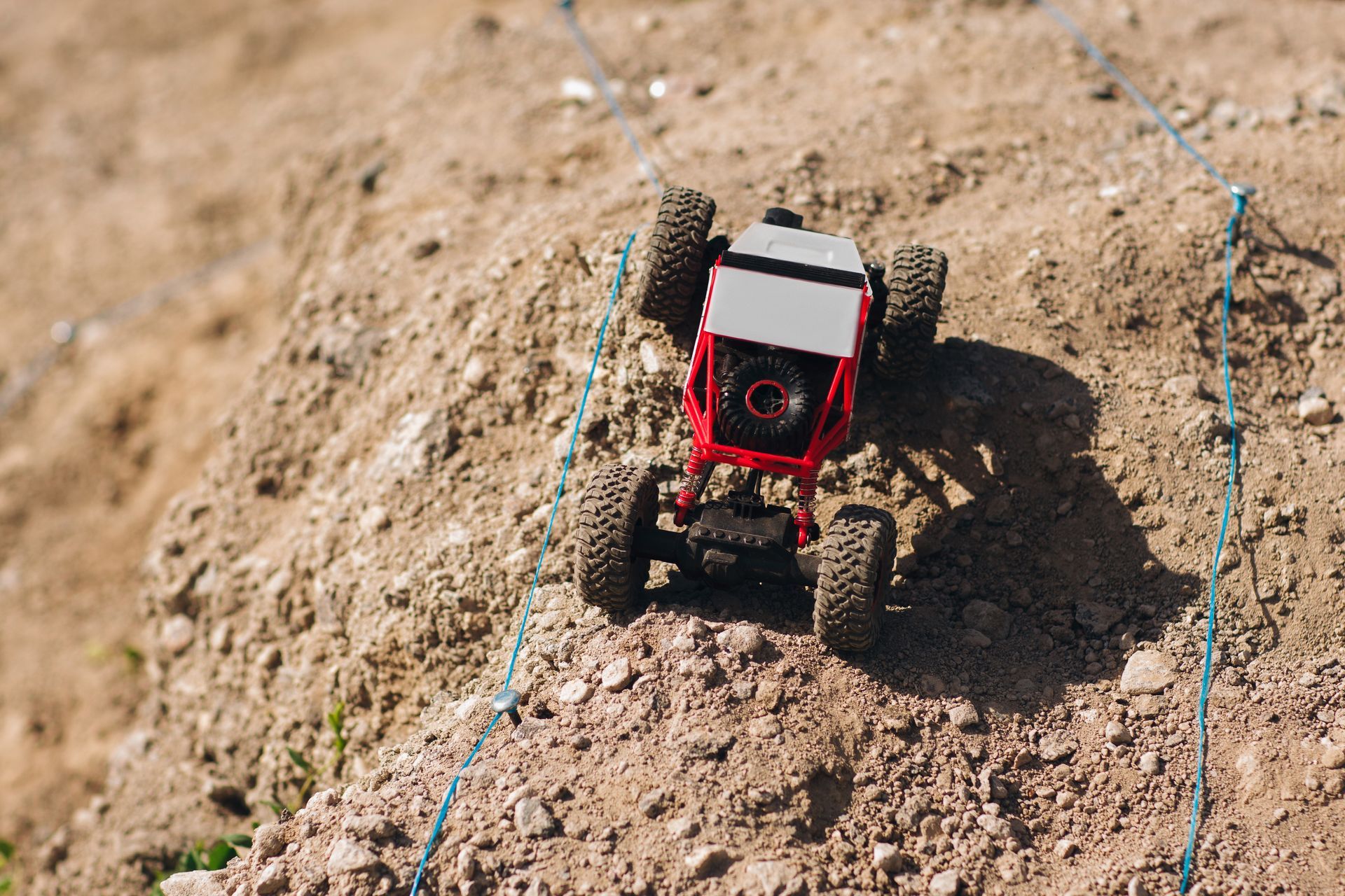 Red and grey remote-controlled car climbing a dirt hill. Blue guide ropes are visible.