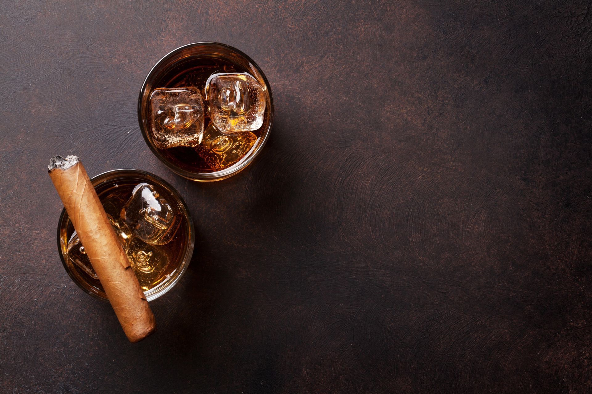Due bicchieri di whisky con ghiaccio e un sigaro su una superficie marrone scuro.