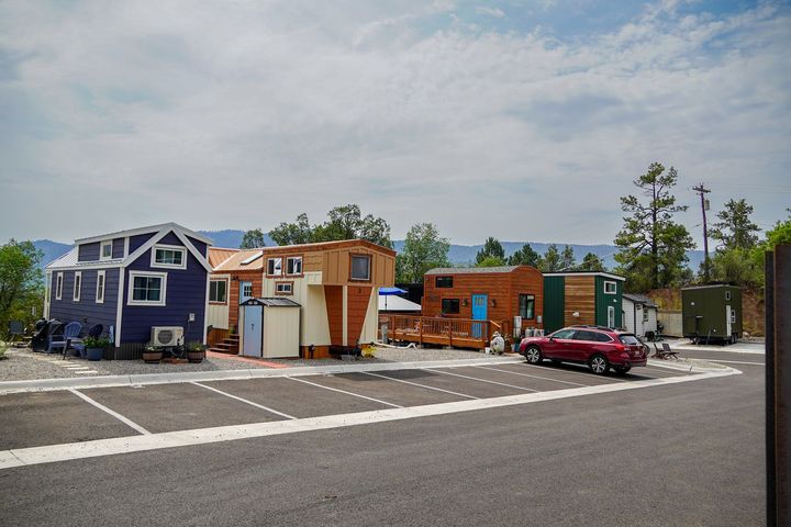 A row of colorful tiny houses with a parking lot in front, against a backdrop of trees and mountains.