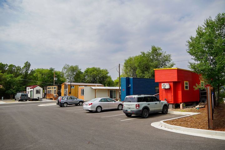 A parking area with several colorful tiny homes and cars parked in front on a cloudy day.