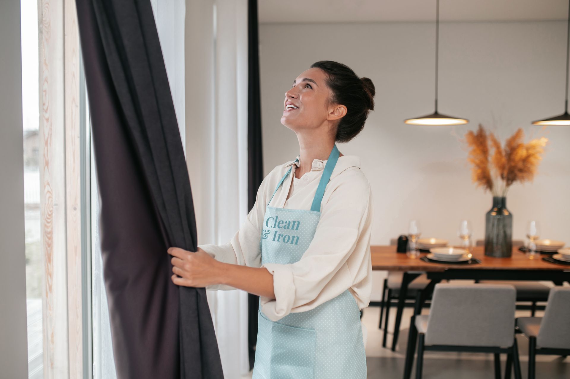Mujer con delantal abriendo cortinas oscuras, sonriendo a la luz del sol. Comedor moderno.
