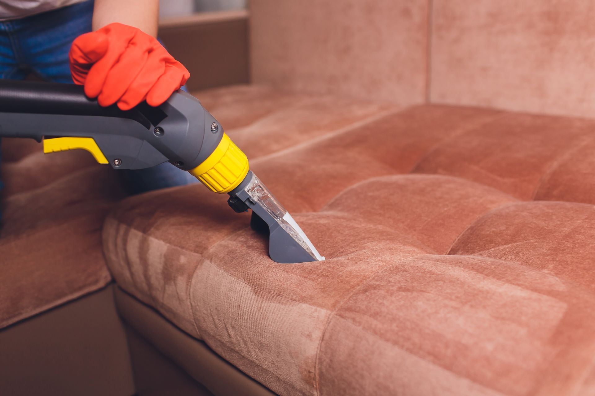 Persona con guantes rojos usando un limpiador de alfombras en un sofá seccional marrón, interior de una casa.