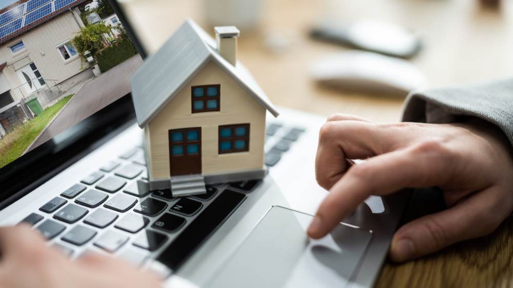 Person using a laptop, a model house sits on it, website with house image on screen.