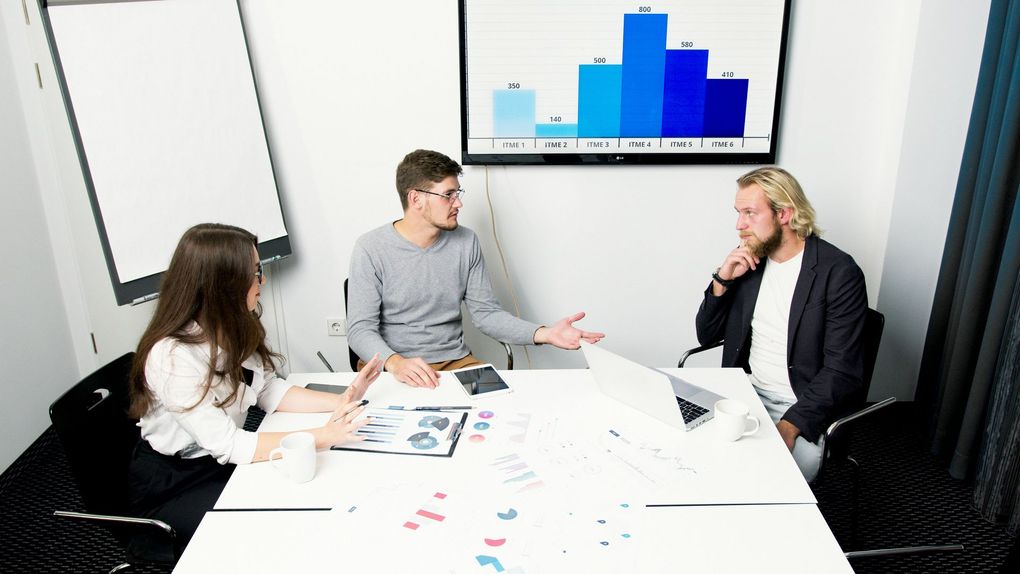 Three people in a meeting, discussing data on a screen and papers, in an office.