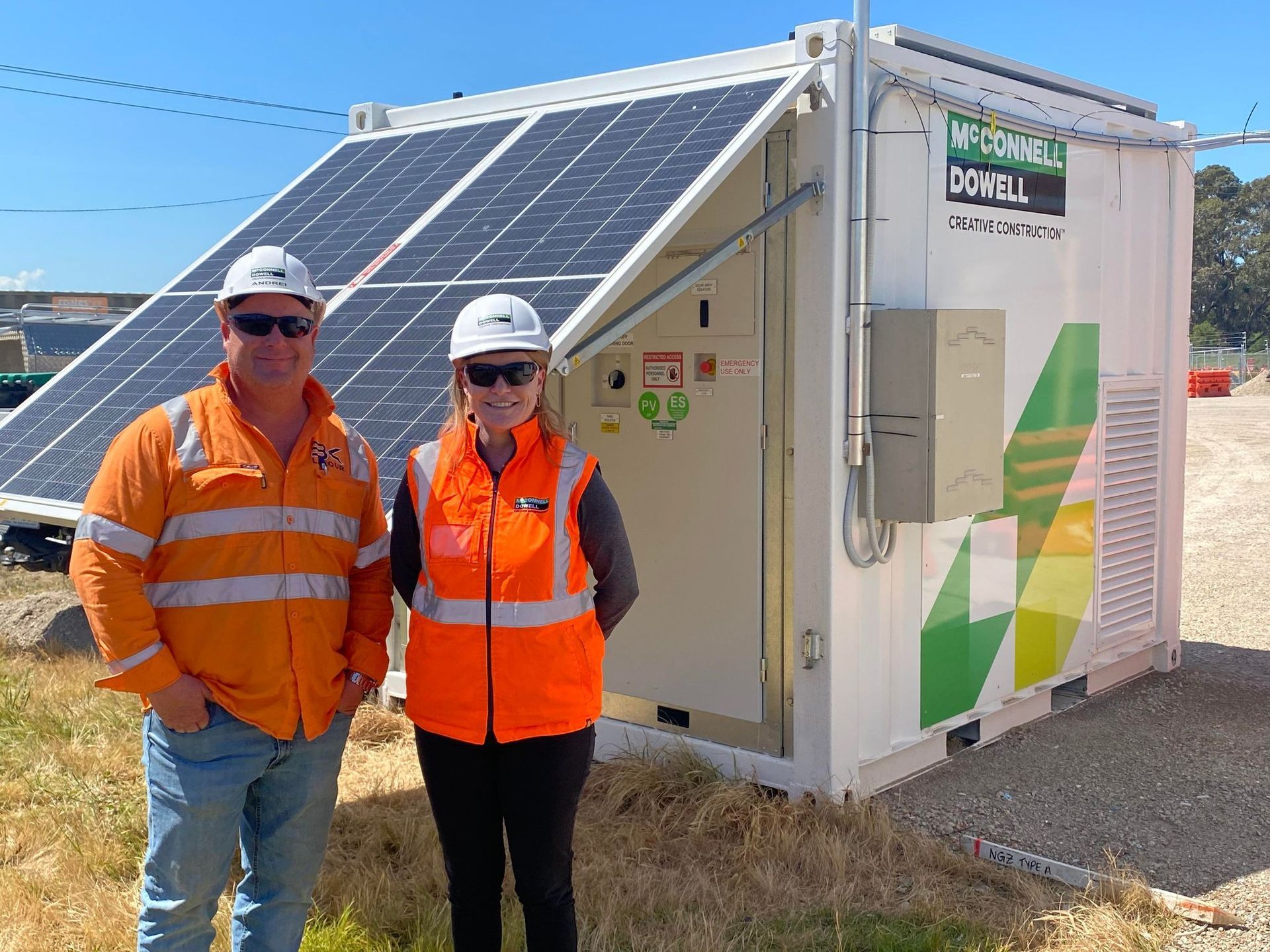 Two people in safety vests stand beside a container with solar panels, outdoors.