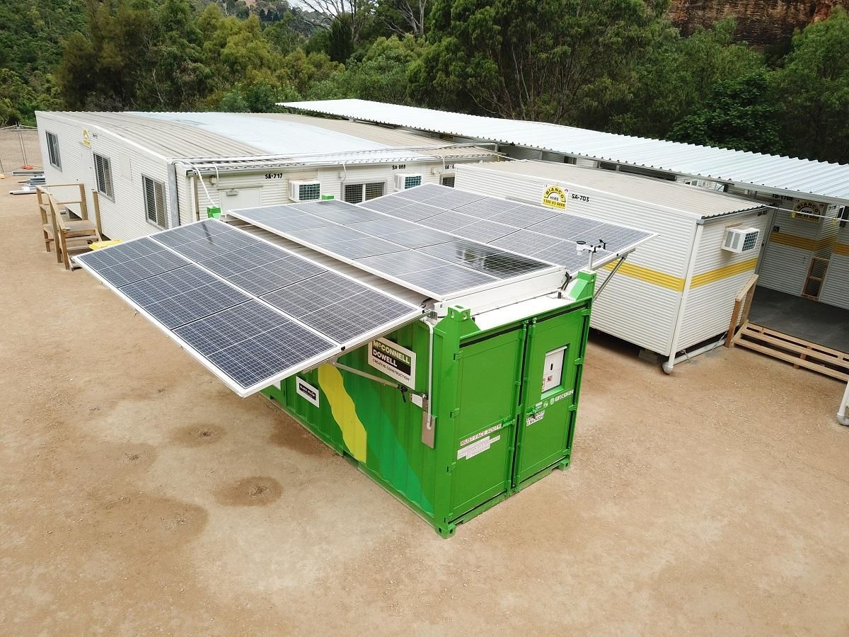 Green container with solar panels open, in front of buildings with solar panels. Outdoors.